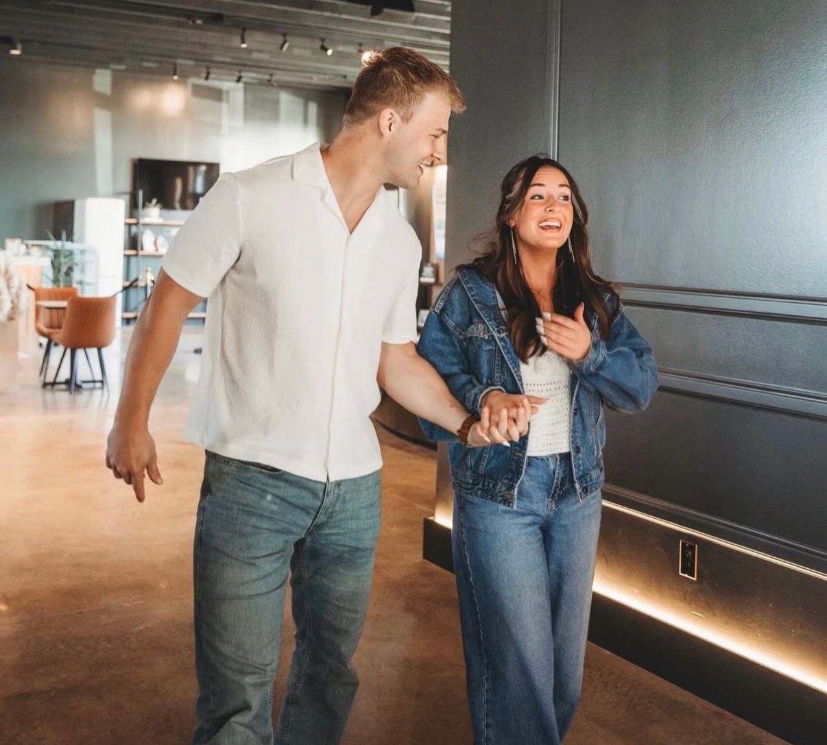 Two people walking and smiling in a modern hallway, holding hands and chatting.