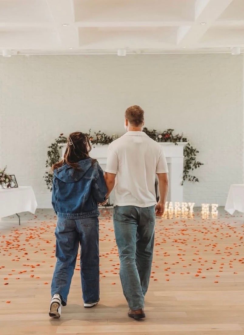 Two people walking down a rose-petal aisle toward a decorated white arch in a bright room