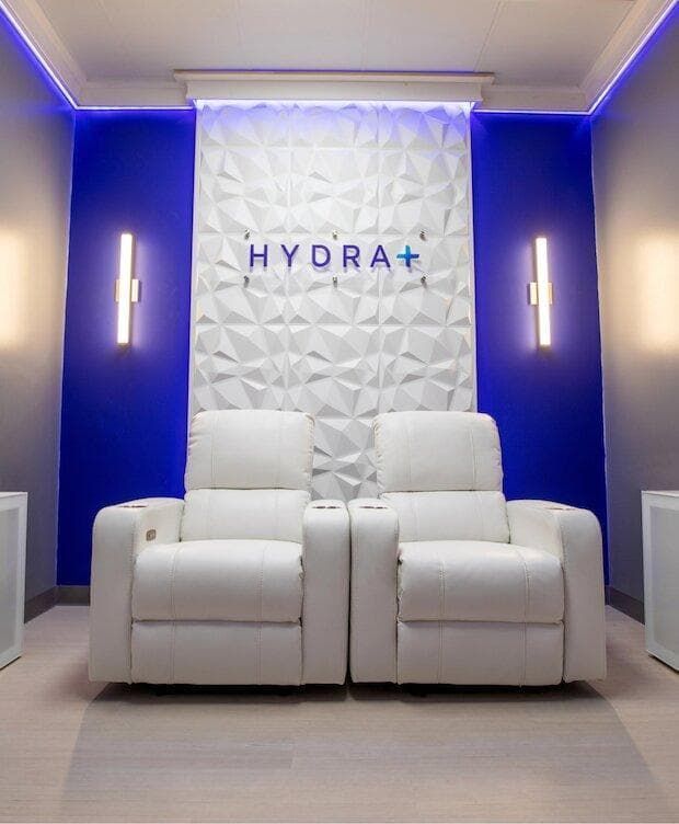 Two white recliner chairs in a blue-lit room with a textured white wall, HYDRA+ logo, and sconces.