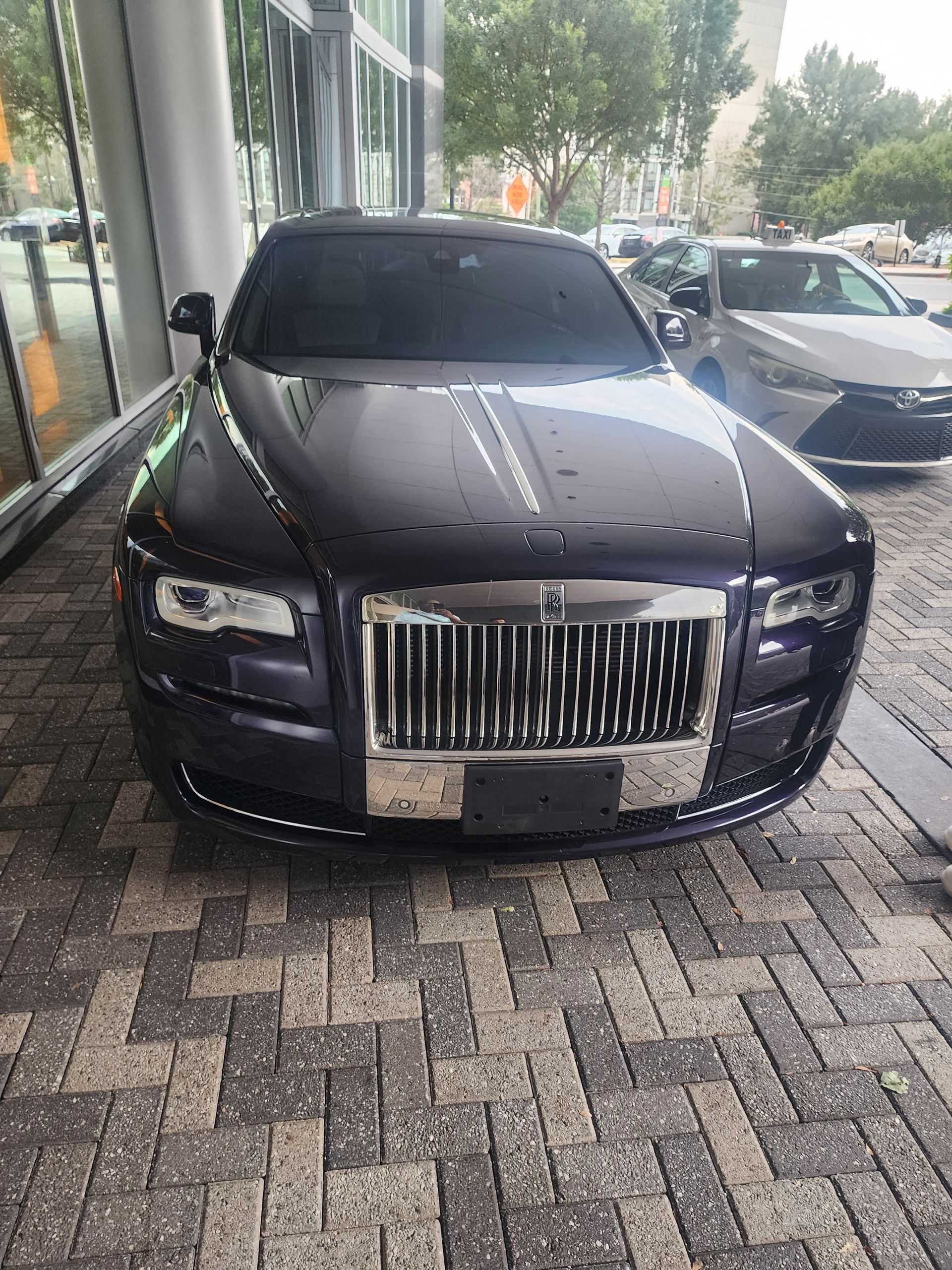 A dark purple Rolls-Royce parked on brick pavement in front of a modern building.