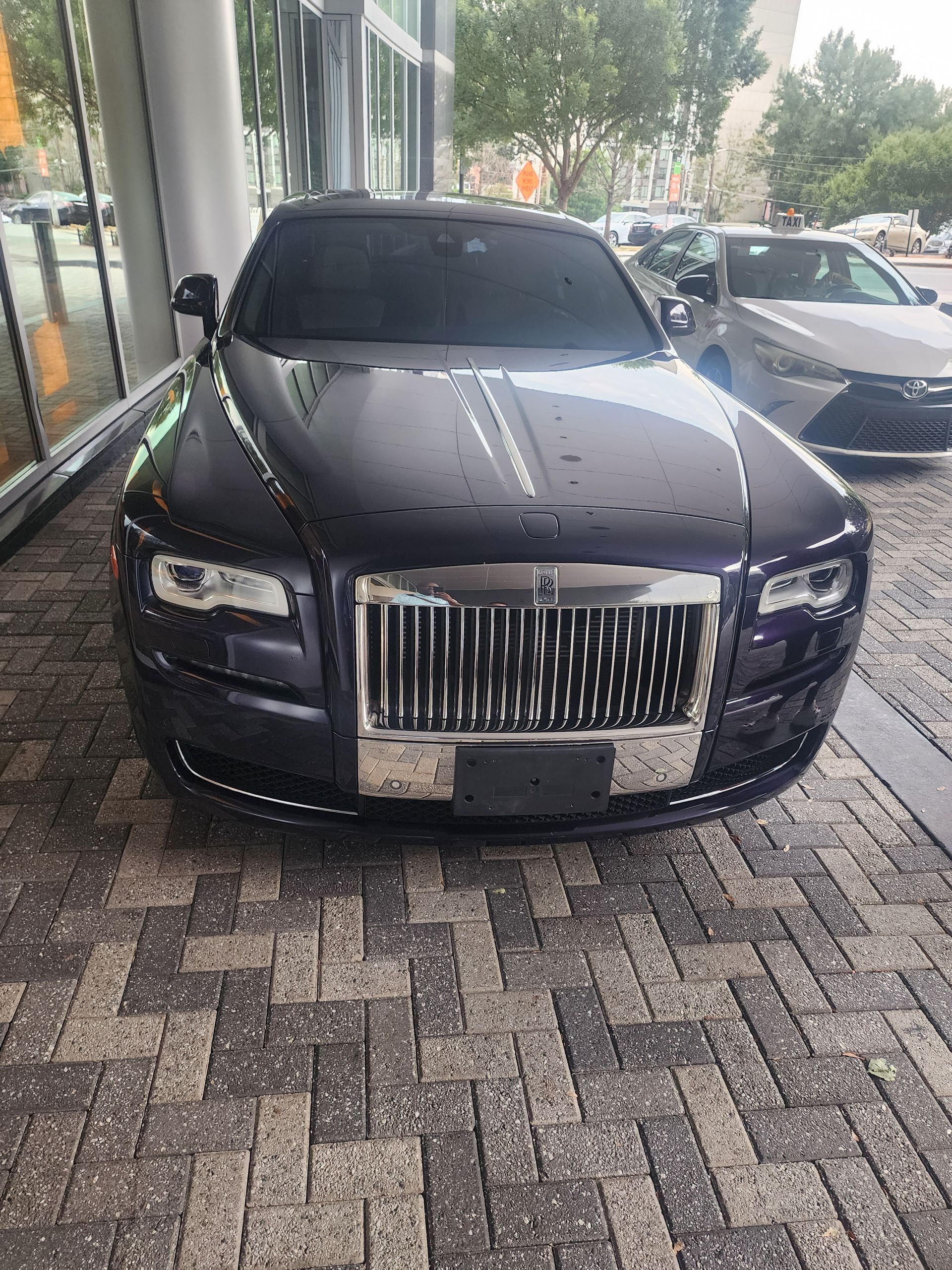 Dark purple Rolls-Royce sedan parked on a brick-paved sidewalk in front of a building with glass windows.