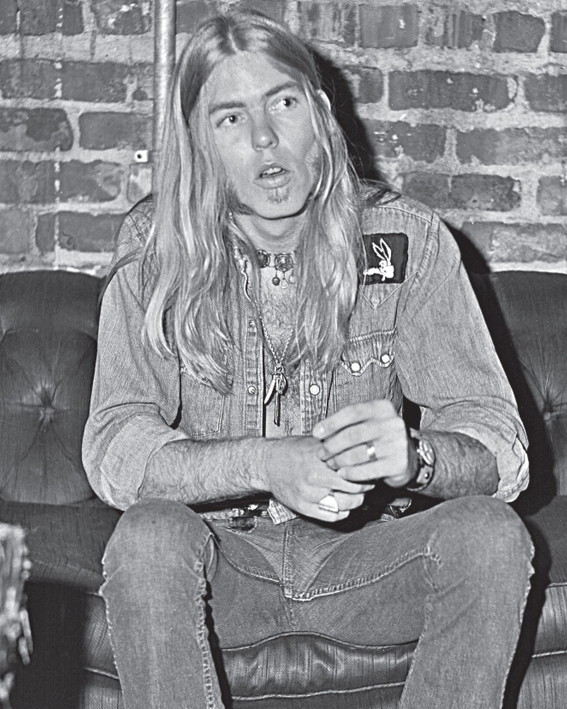 Long-haired person sitting on a couch against a brick wall, holding a cup and speaking.