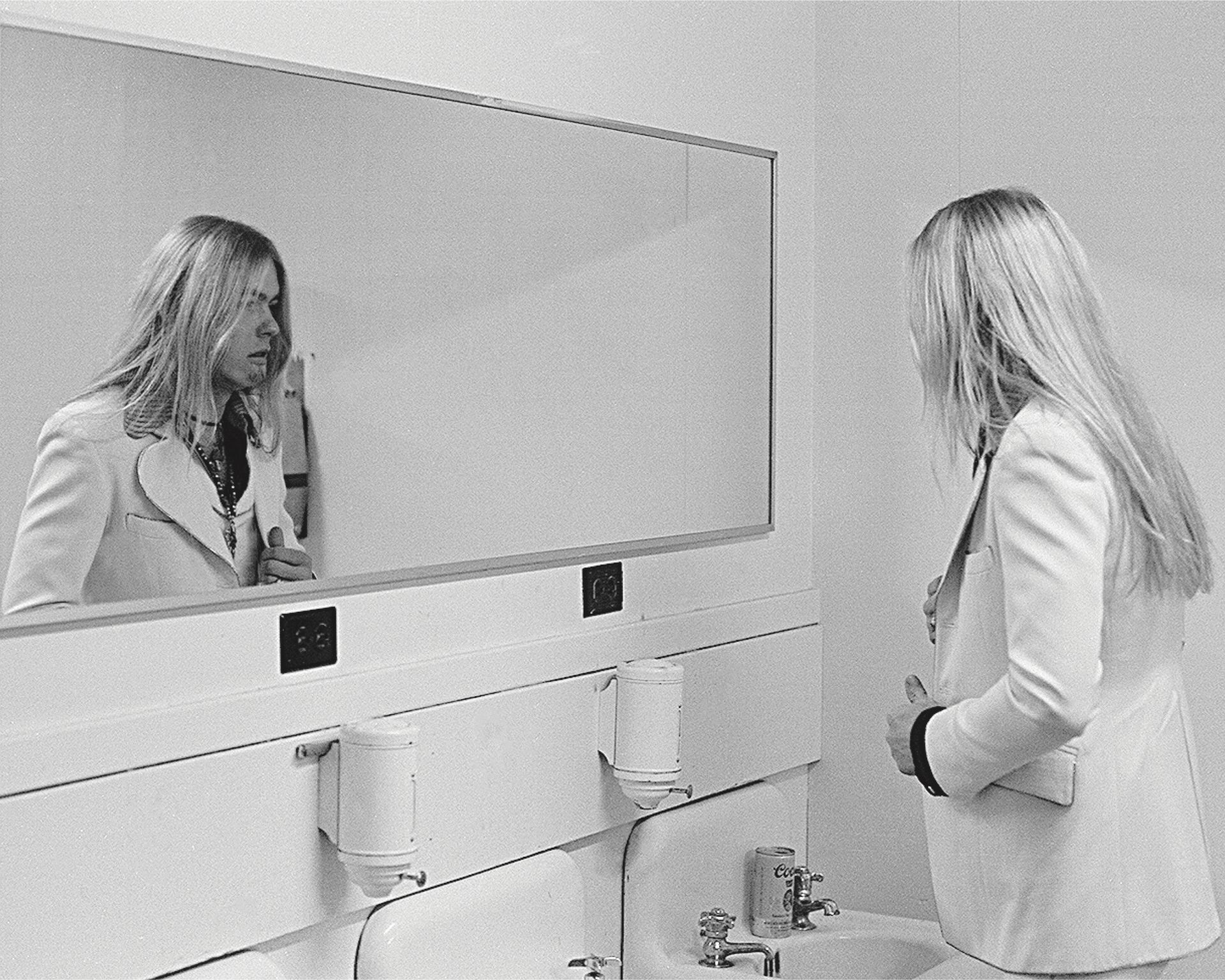 Person looking in a bathroom mirror at a sink and soap dispensers in a monochrome restroom