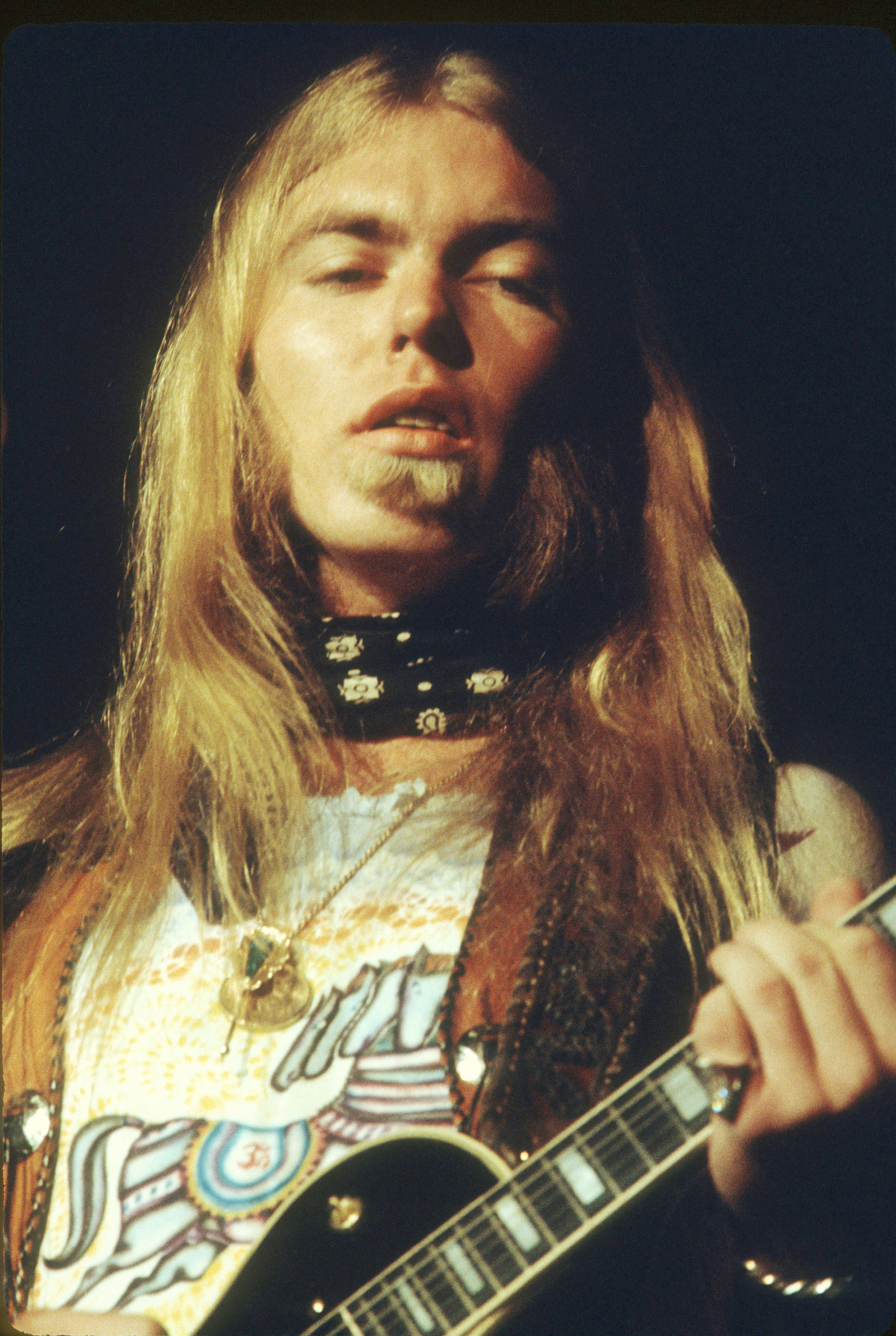 Blond musician with eyes closed, holding a black electric guitar on a dark stage.