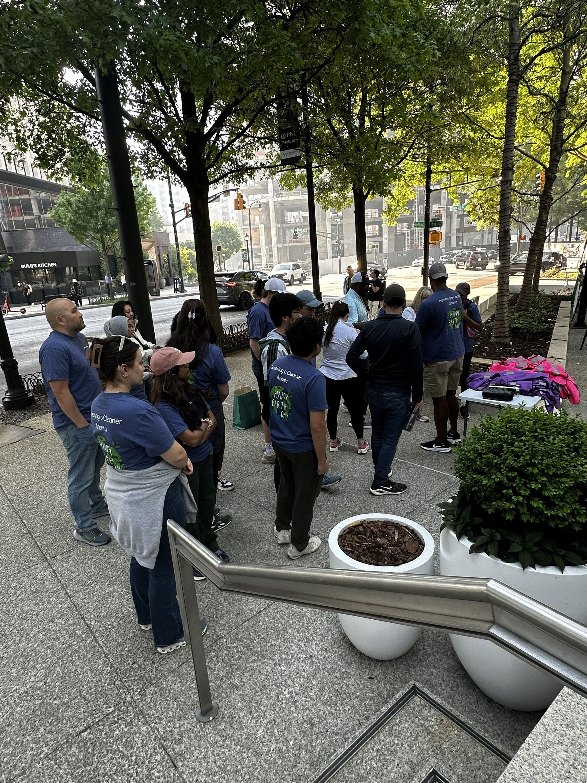 People gathered in a tree-lined plaza near a stairway and large white planters.