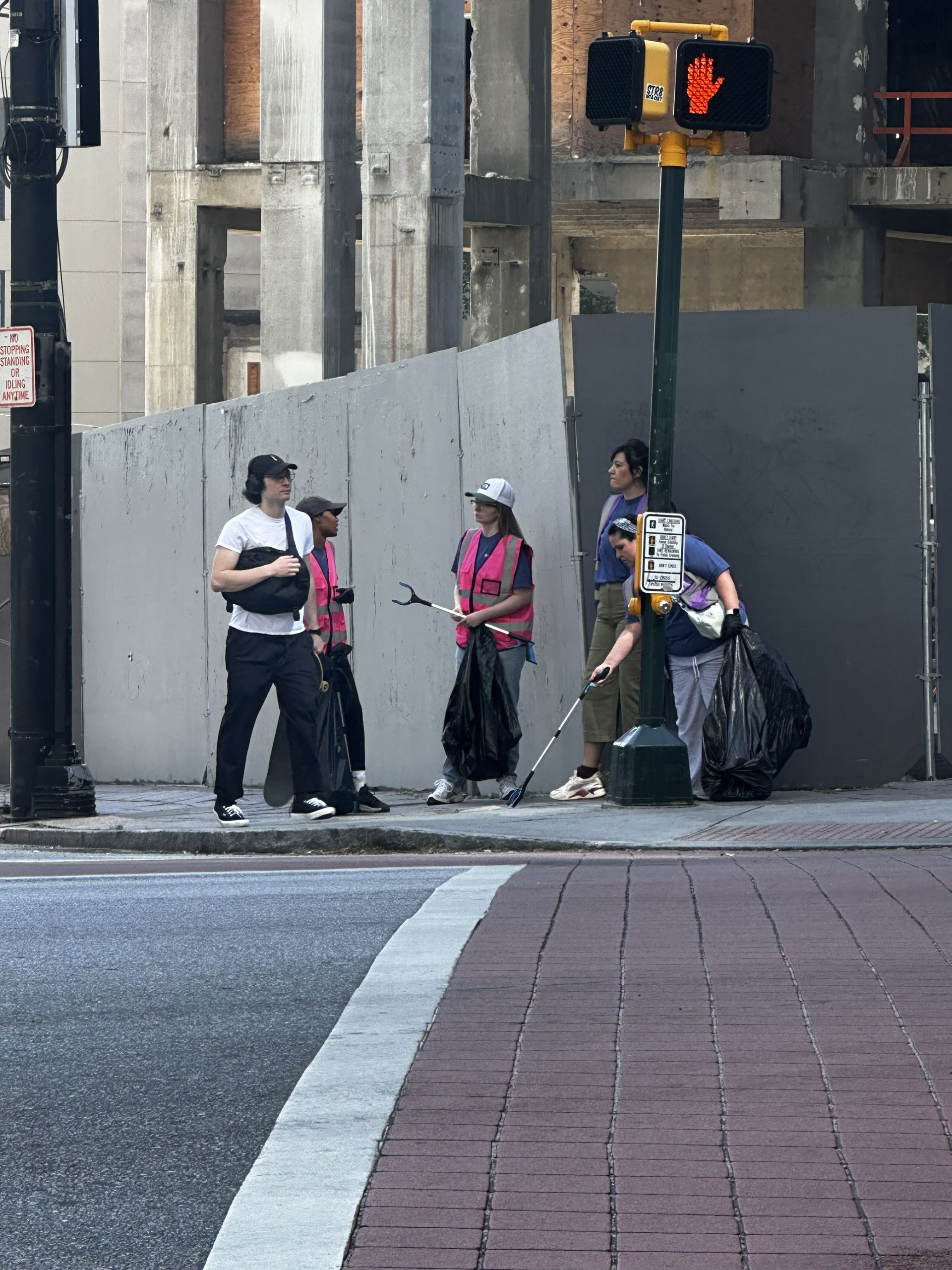 Pedestrians crossing at a city intersection beside a crosswalk and traffic light, with one person sweeping debris.