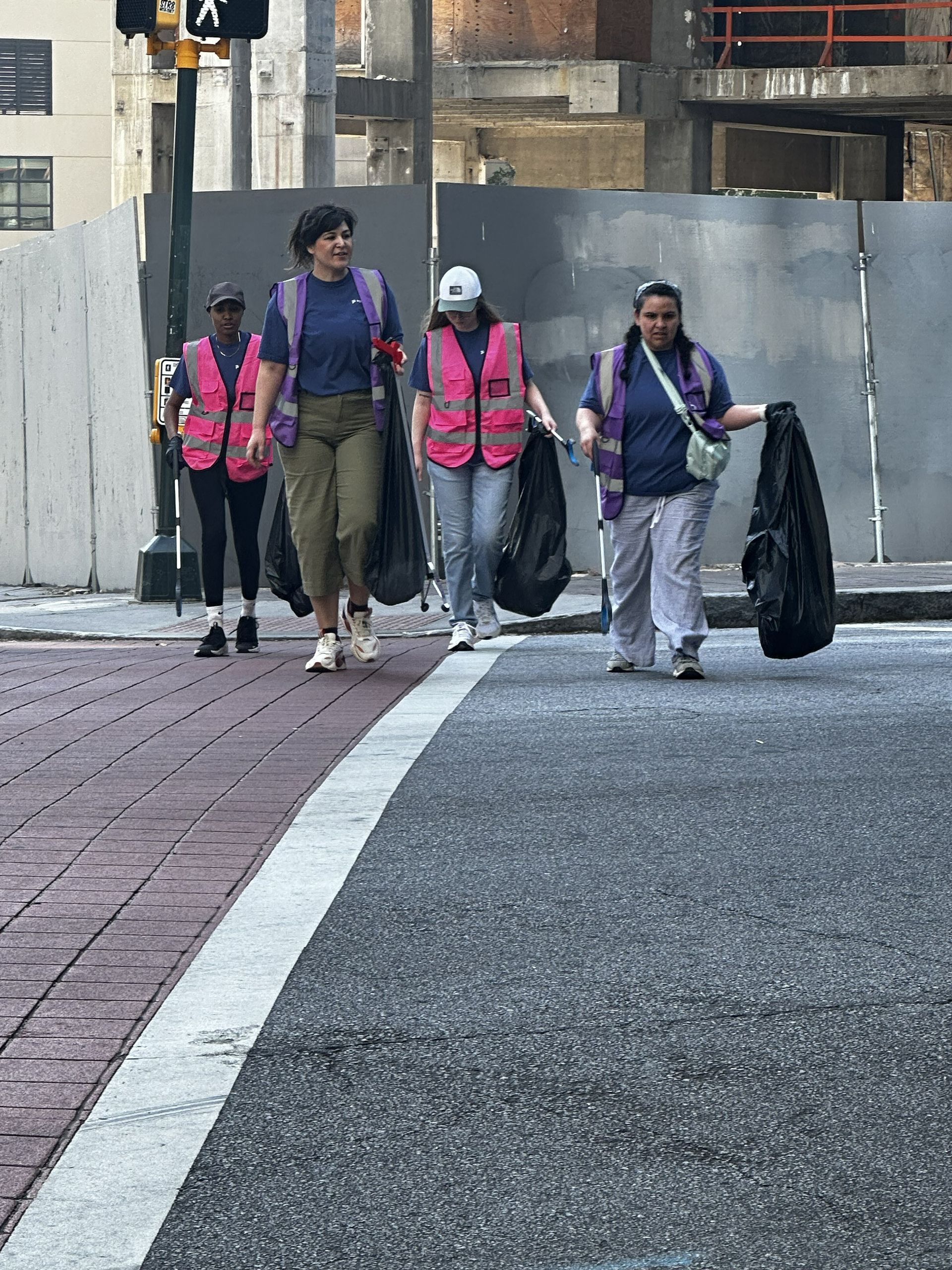 Four pedestrians in bright pink safety vests walking along a city sidewalk near a construction site