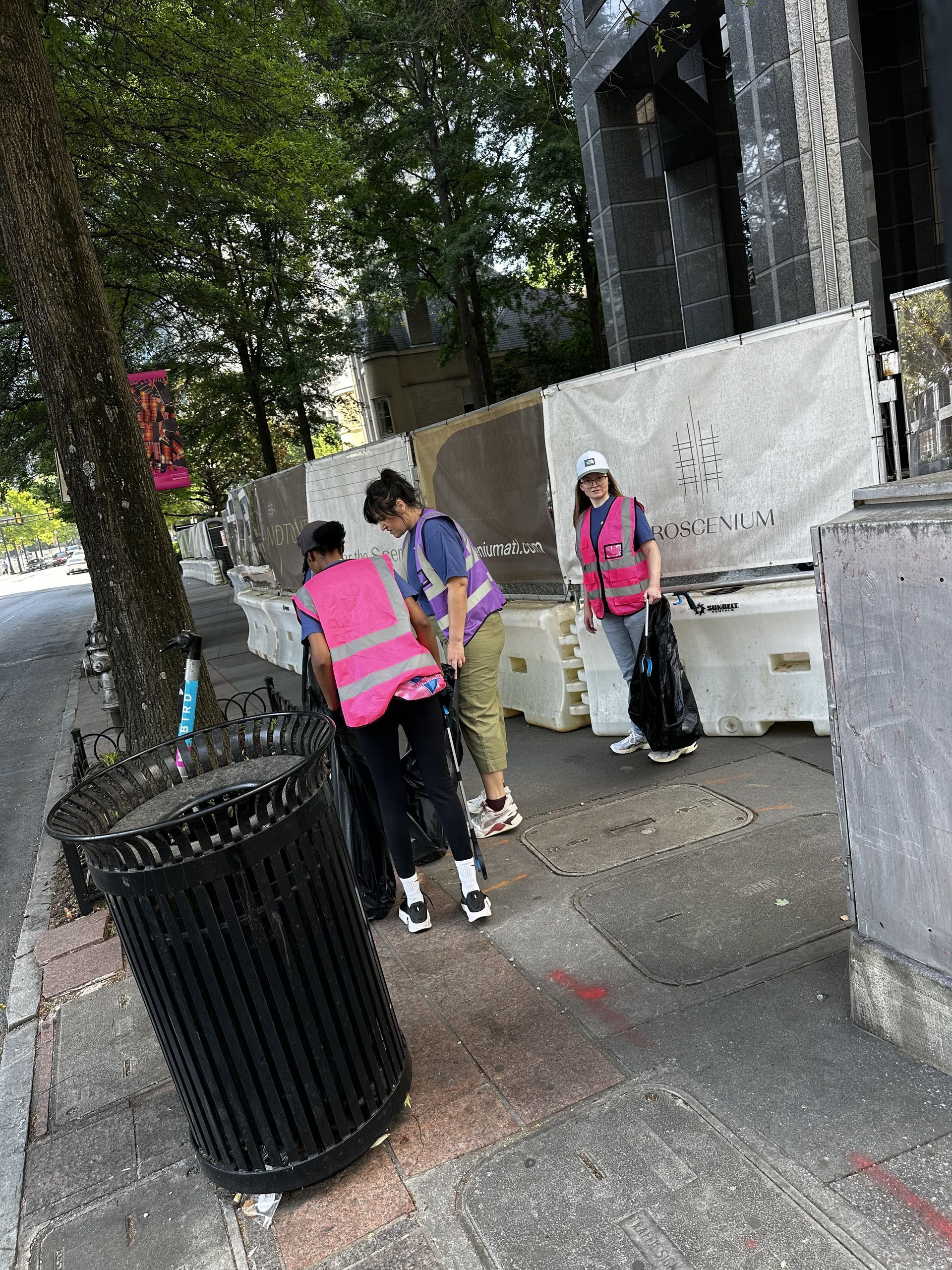 People in pink vests standing on a sidewalk near a trash can and a tree by a building.