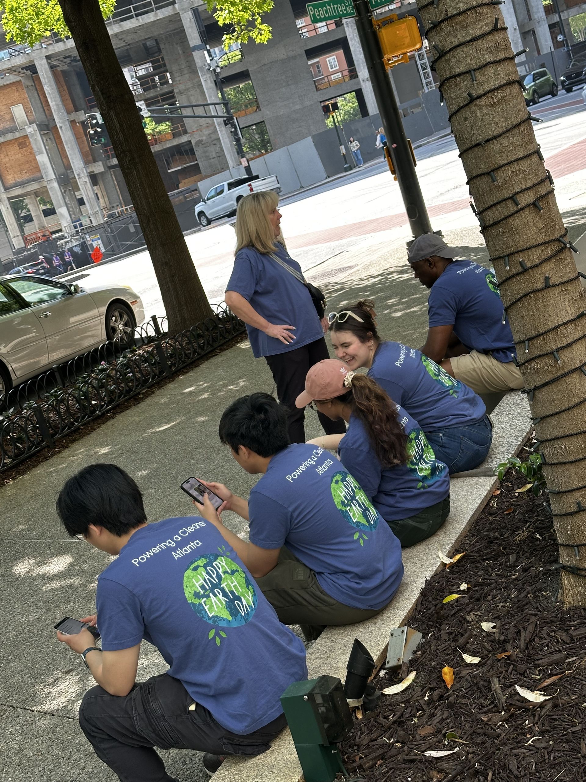 People in blue shirts sitting on a sidewalk under a tree, looking toward a street with parked cars.