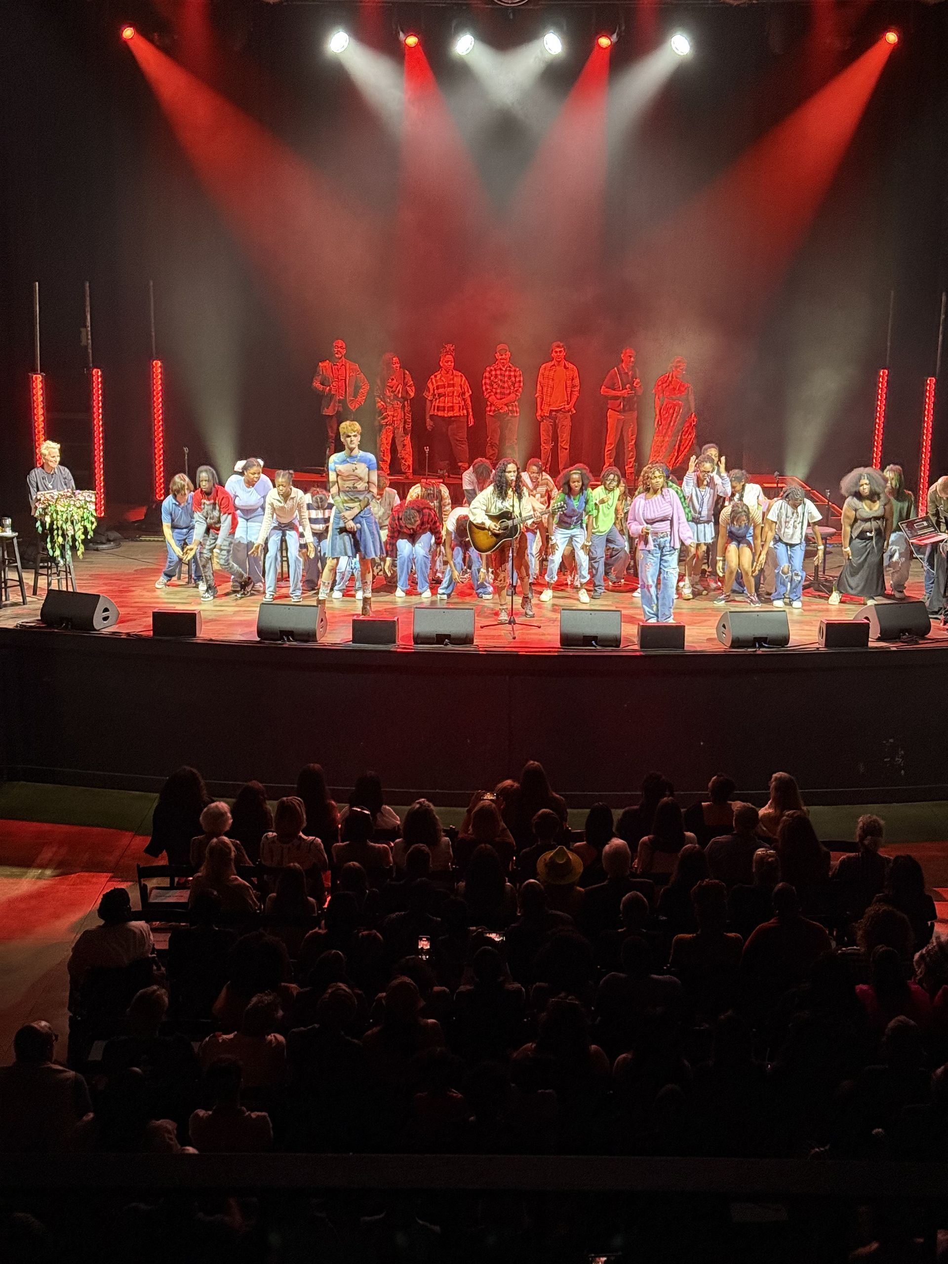 A group of people on a stage with a band, red lights and an audience.