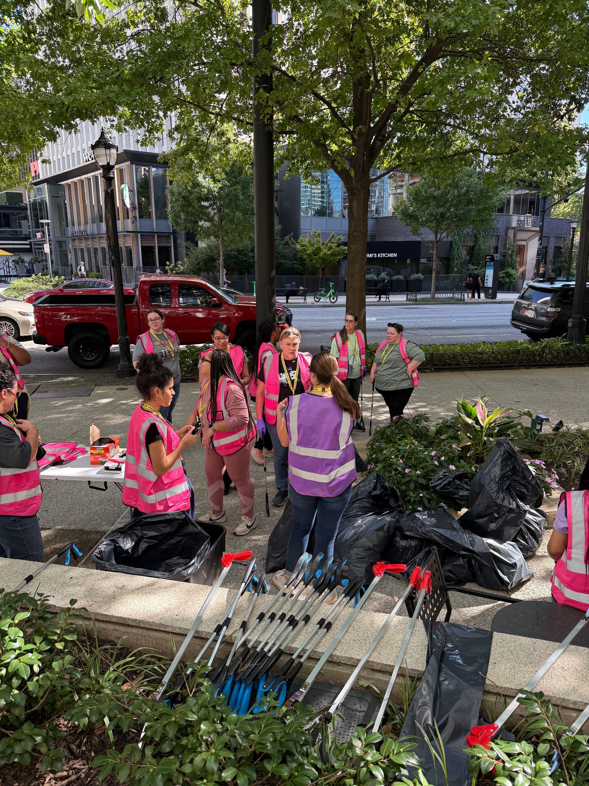 Group of people in pink vests cleaning a public space, surrounded by trees and trash bags.
