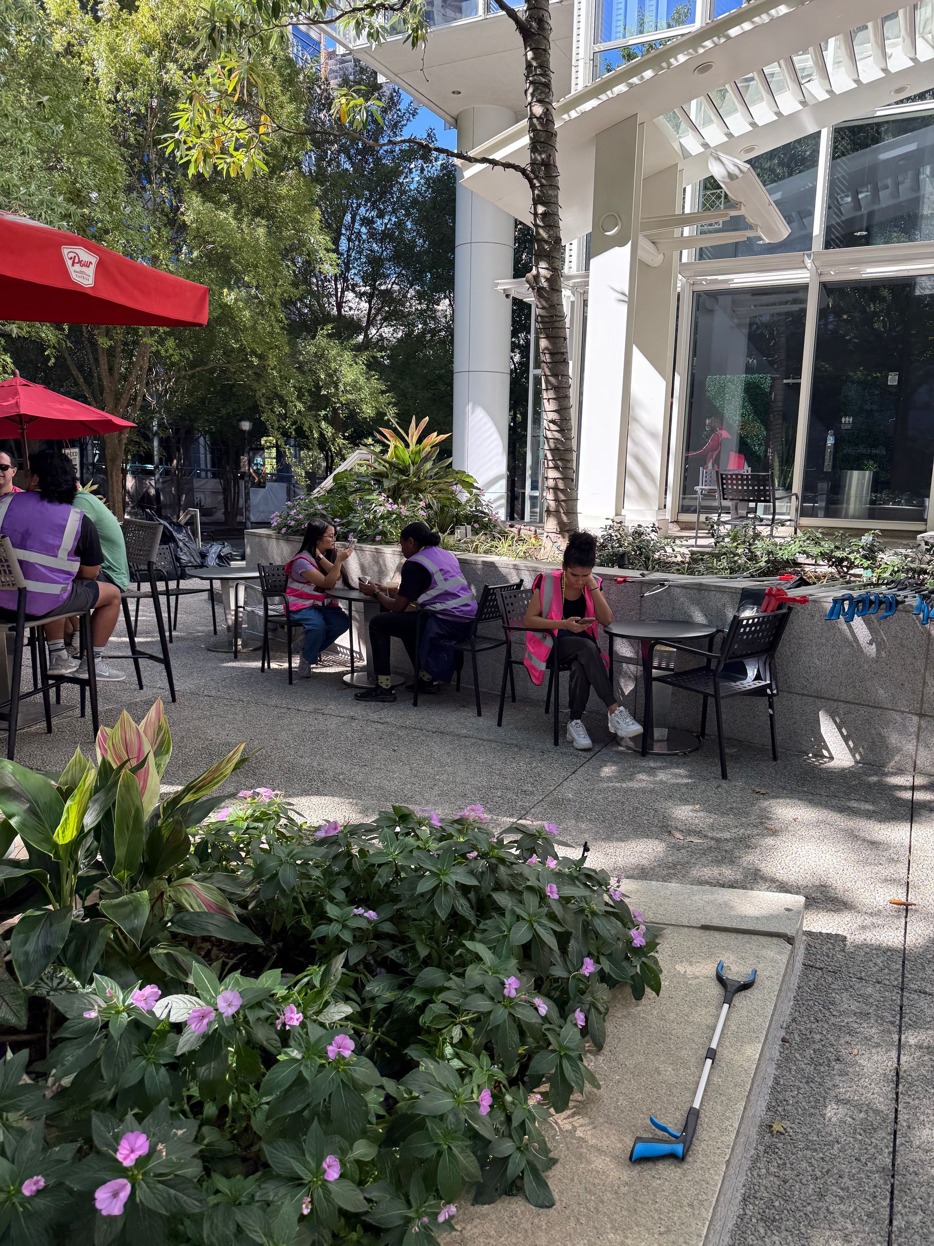 Outdoor cafe with patrons seated at tables; greenery and building in background.