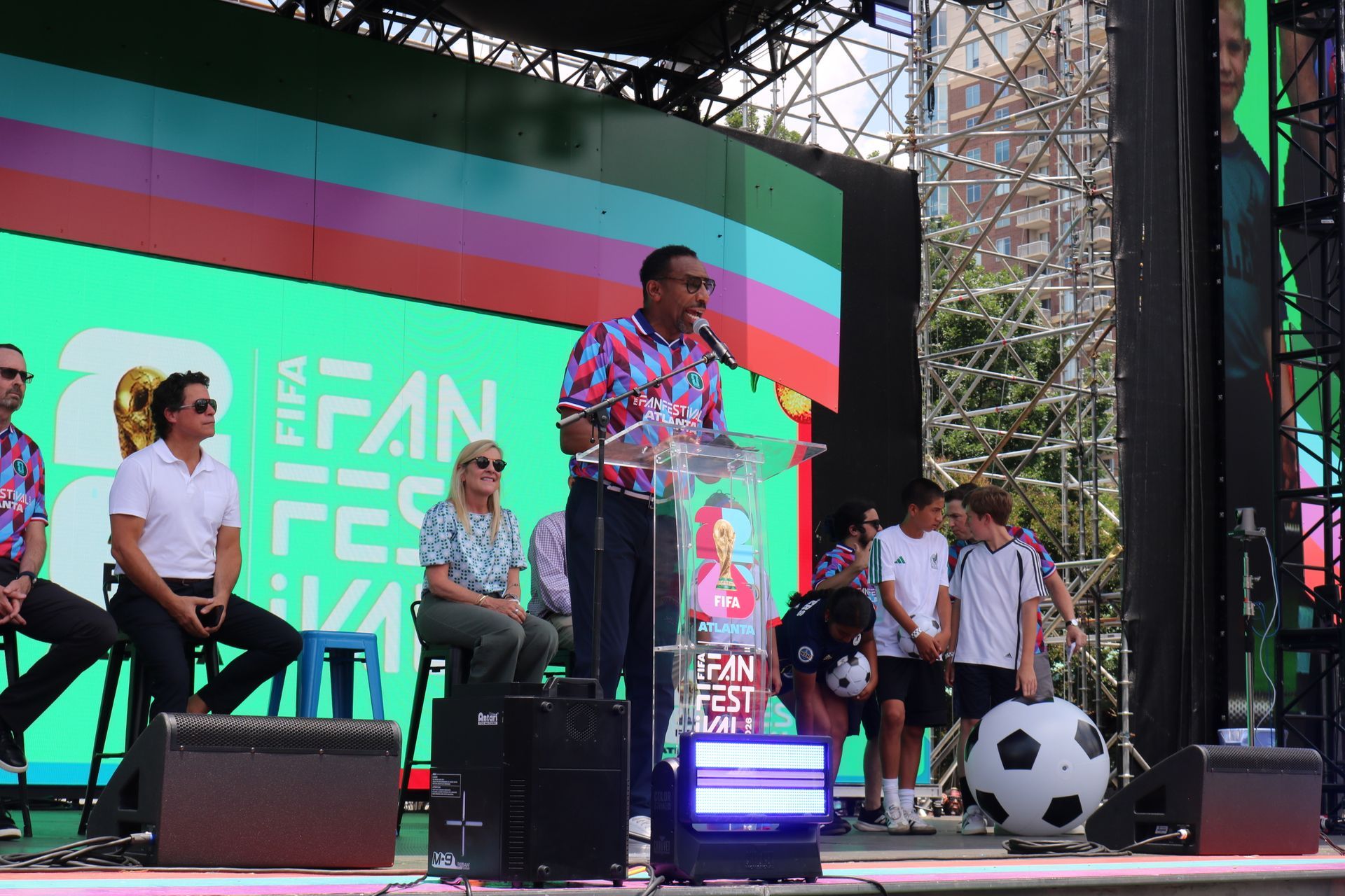 Speaker at podium on outdoor stage with seated panelists and colorful festival backdrop