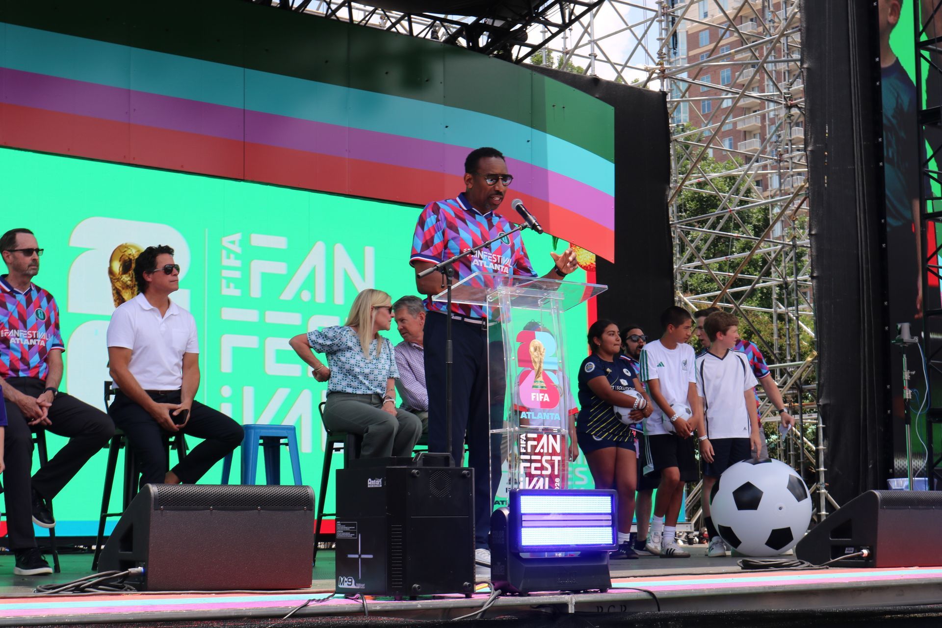 Speaker at podium on outdoor stage with colorful backdrop and people seated nearby