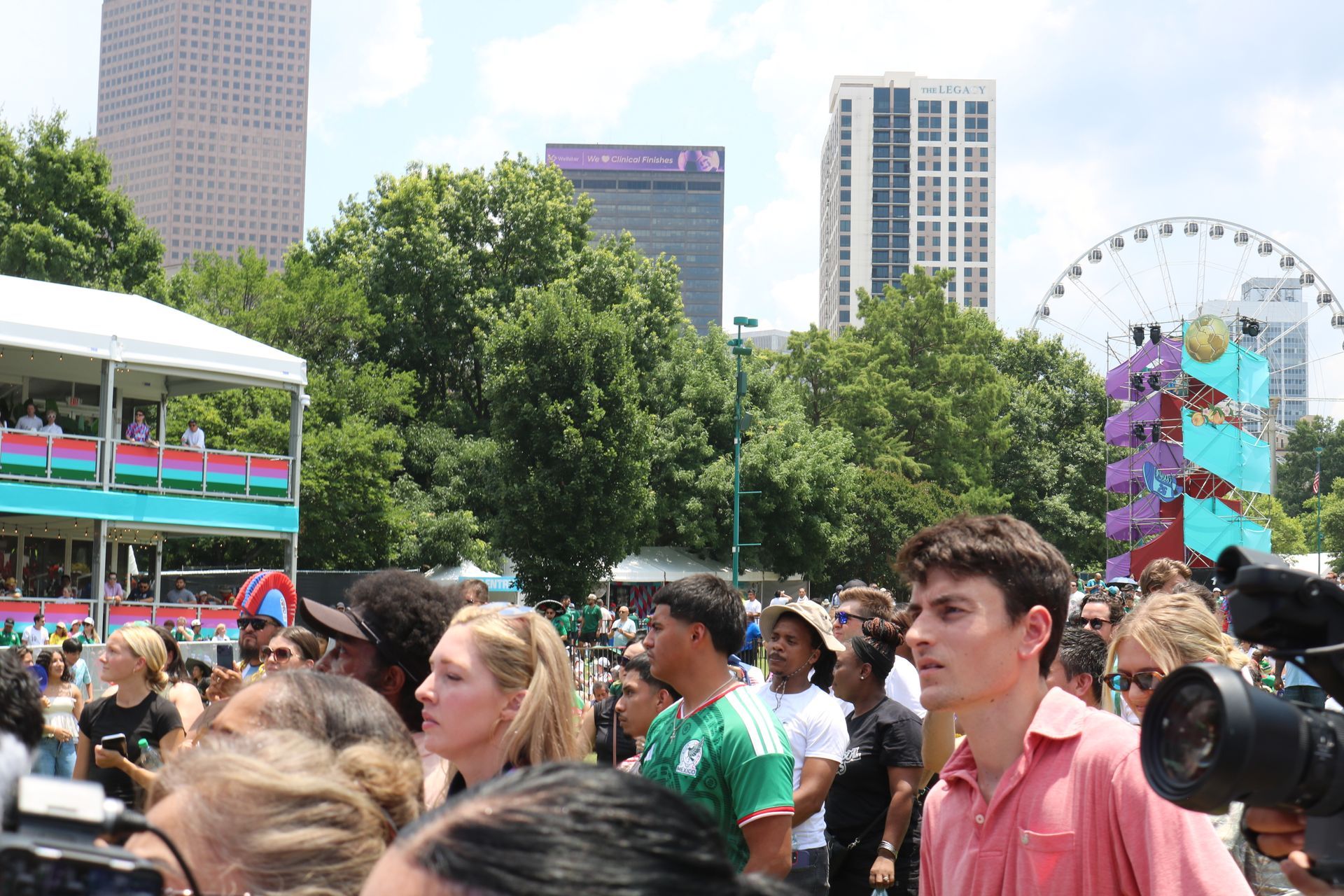 Crowd at a sunny outdoor festival with city buildings and a Ferris wheel in the background