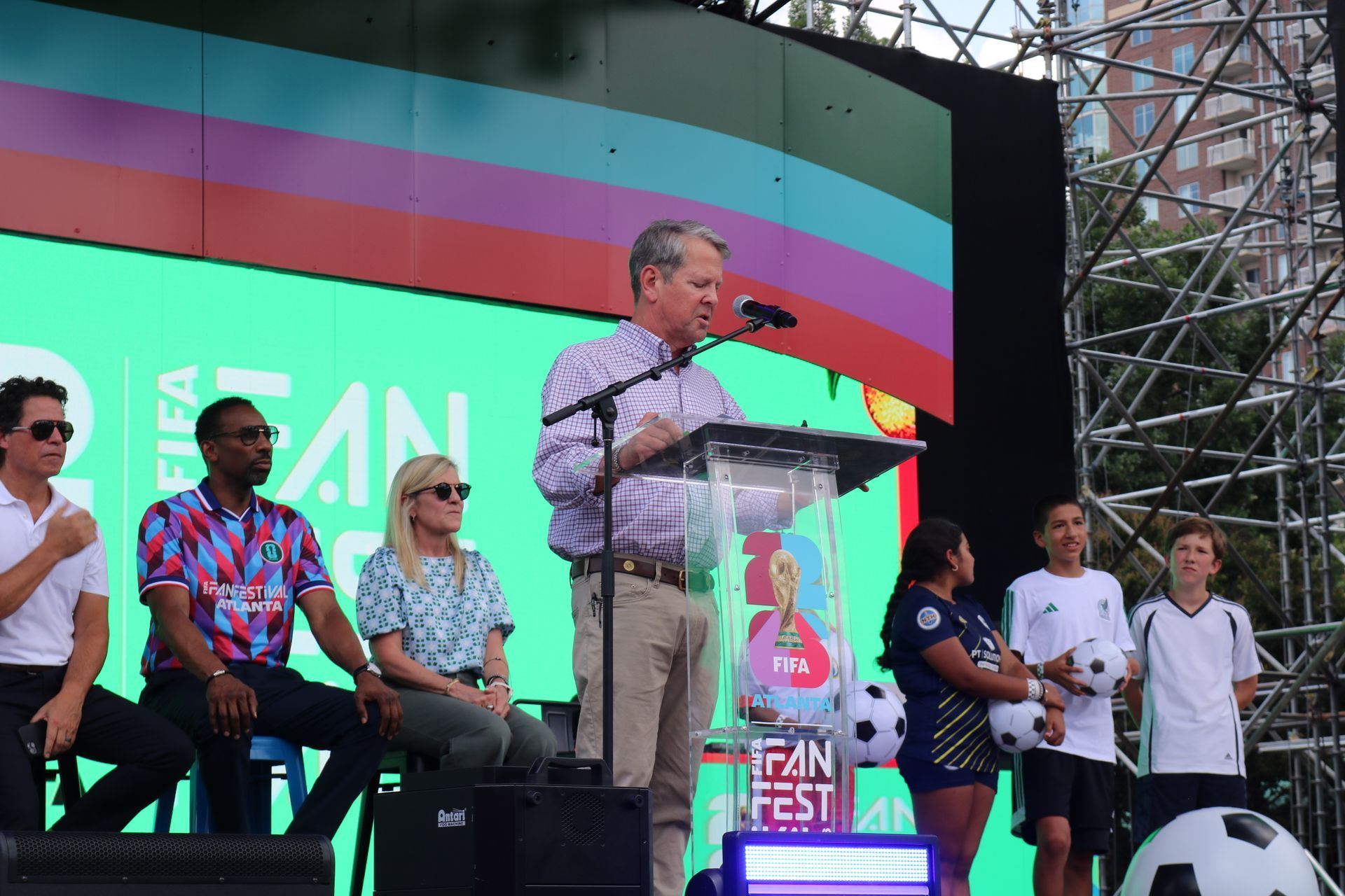 Speaker at podium on outdoor stage with colorful backdrop, seated panelists nearby