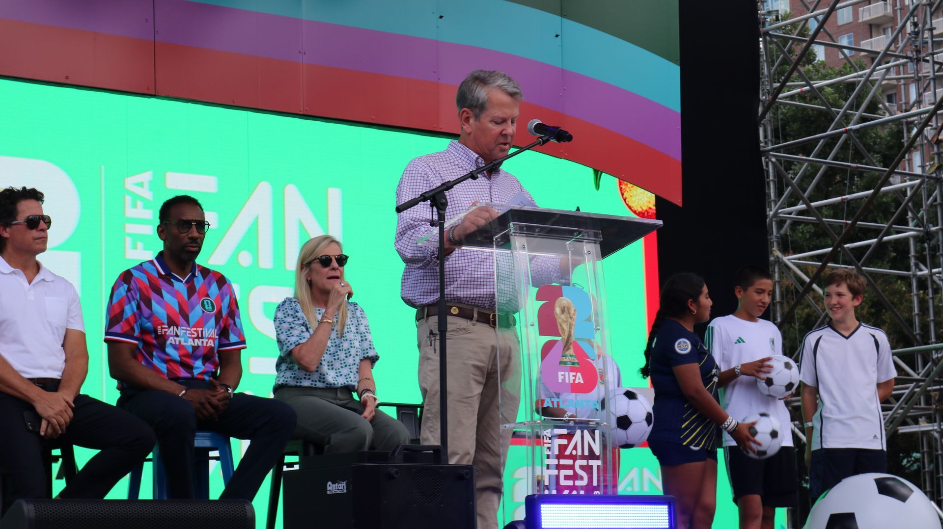 Speaker at podium on stage with seated guests and colorful event backdrop, addressing a crowd