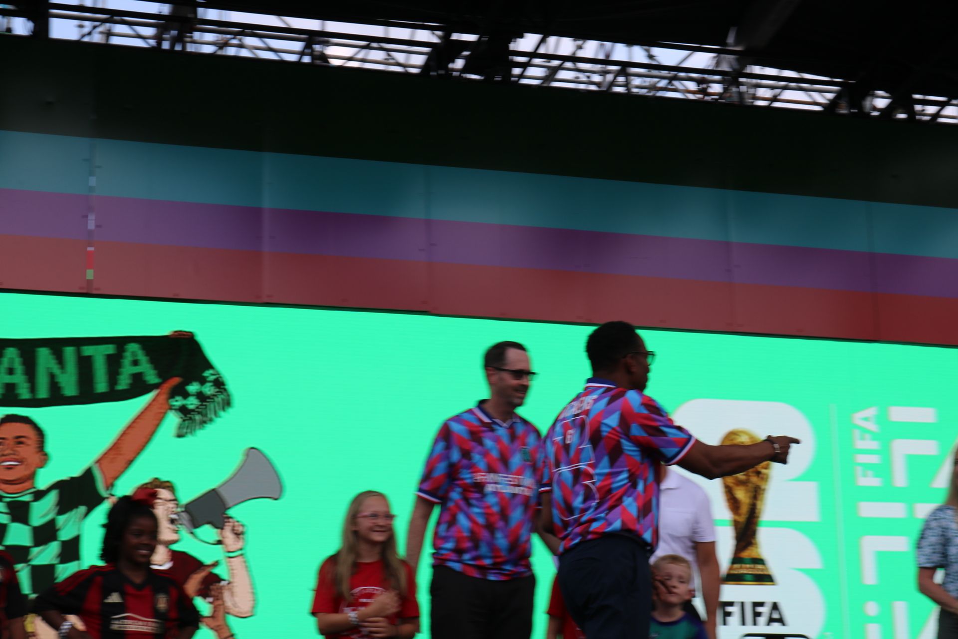 Dancers in colorful patterned shirts perform onstage against a bright green FIFA World Cup backdrop.