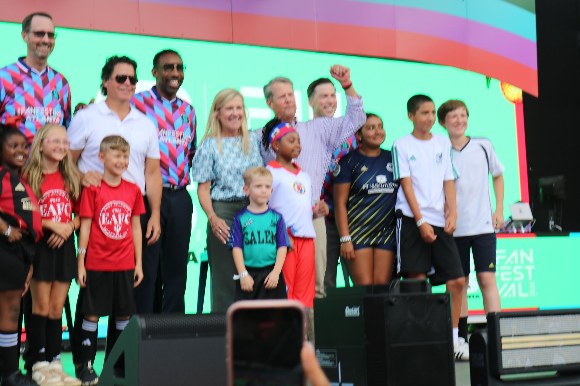 Group of people standing on a stage in colorful shirts, posing for a photo at an outdoor event.