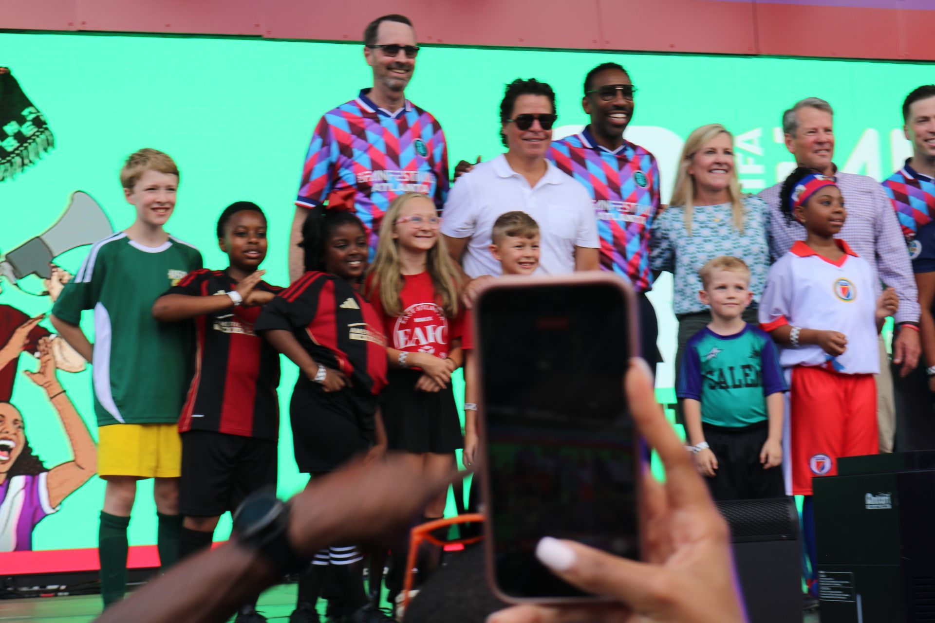 People on a stage posing in front of a bright green screen, with a phone held up in the foreground