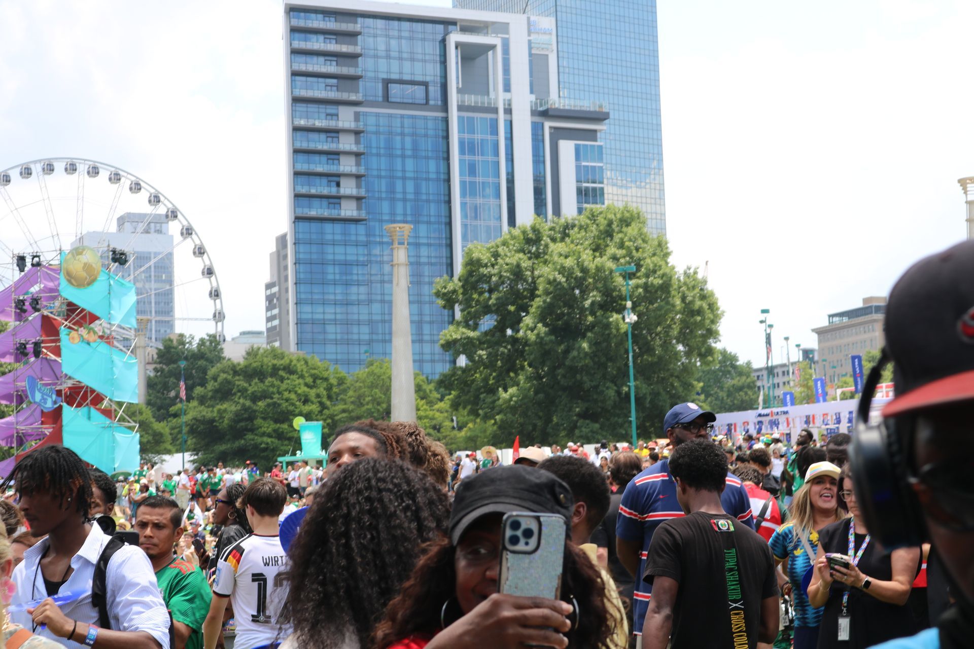 Crowd at outdoor festival near a Ferris wheel and tall blue glass building, with people walking and taking photos