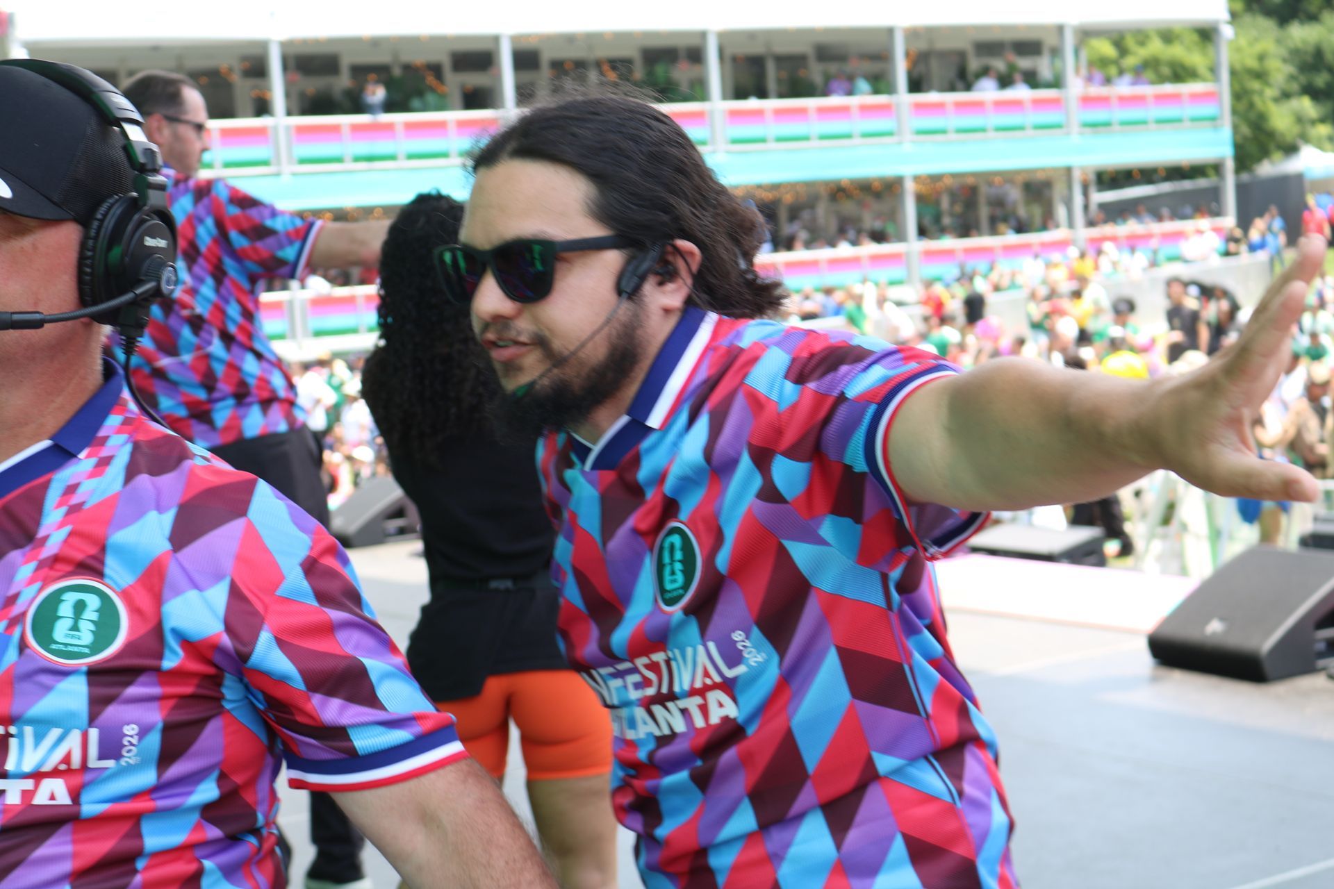 Two men in matching colorful shirts at a stadium, one wearing sunglasses and gesturing with arms outstretched