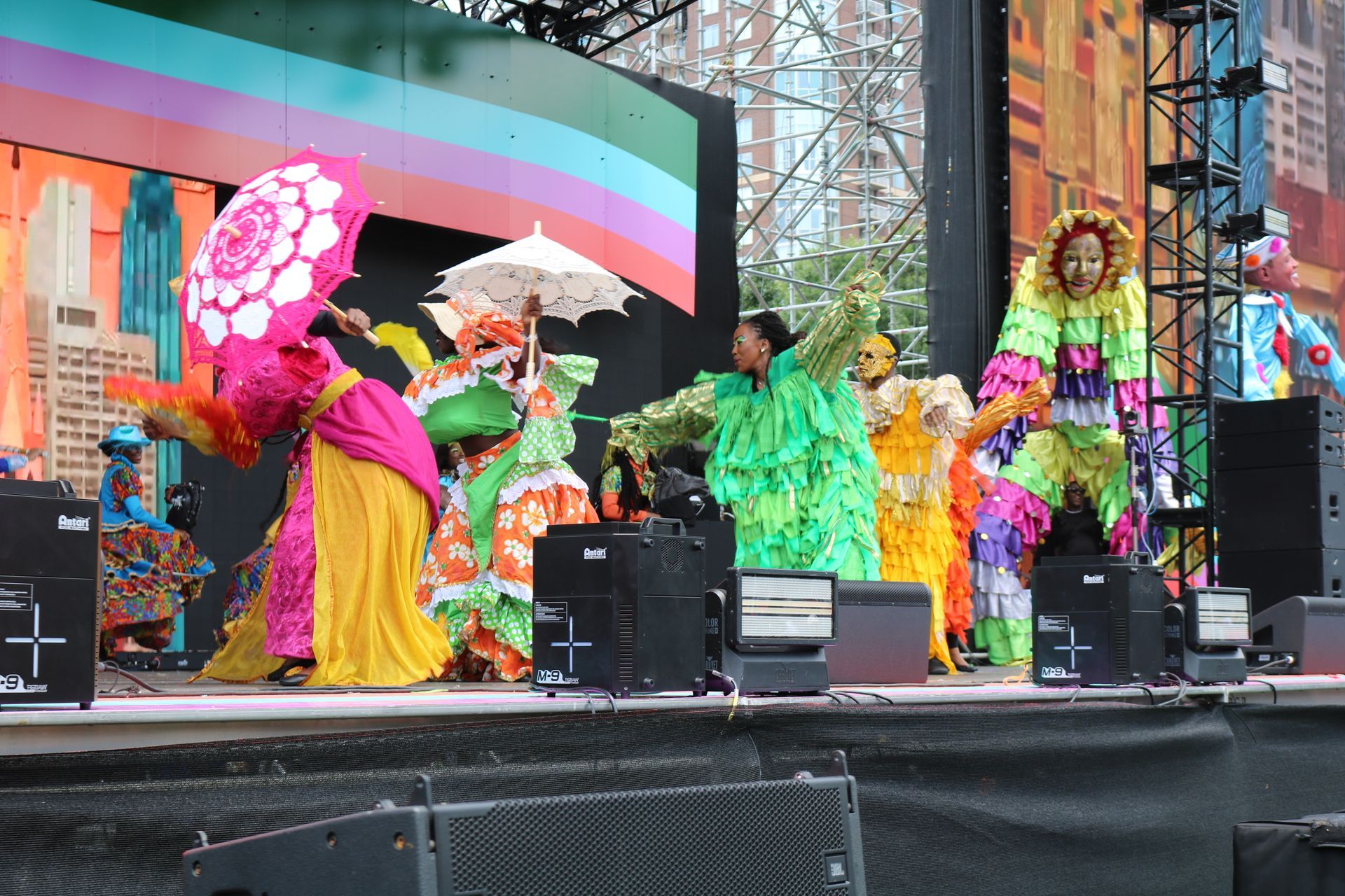 Colorful costumed performers dancing on an outdoor stage with large rainbow and pink props
