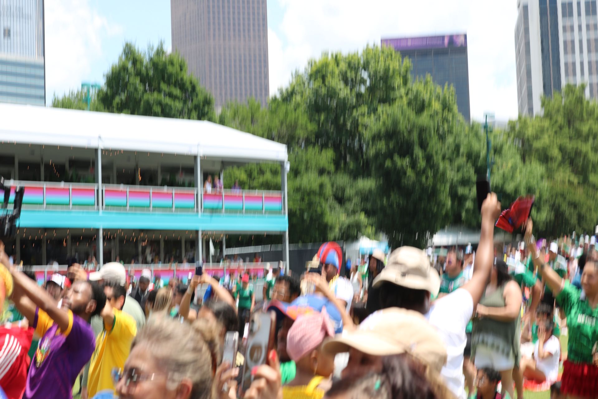 Crowd dancing at an outdoor festival with a stage, trees, and city buildings in the background