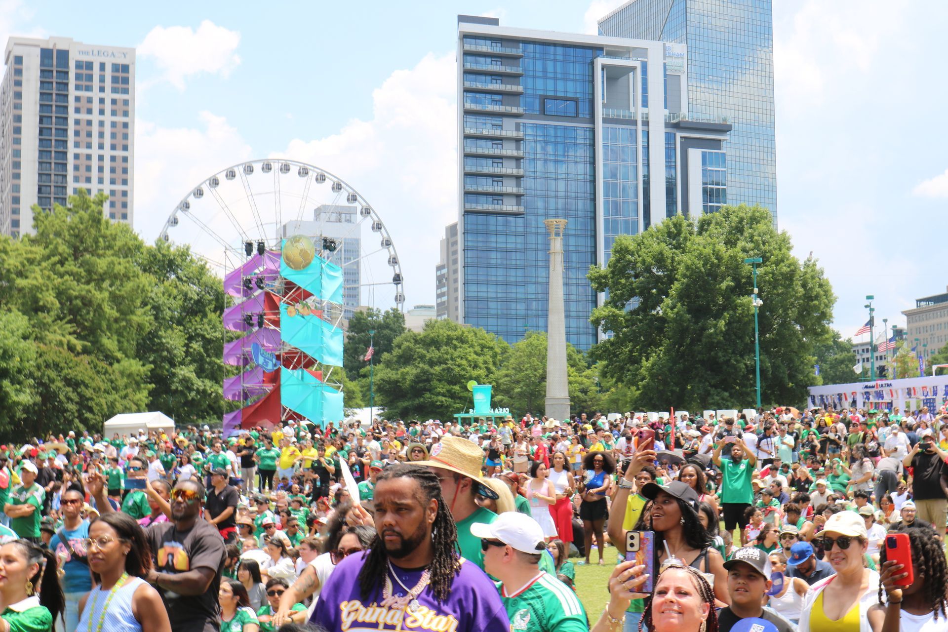 Crowded outdoor festival with colorful Ferris wheel, tall buildings, and people gathered in a park