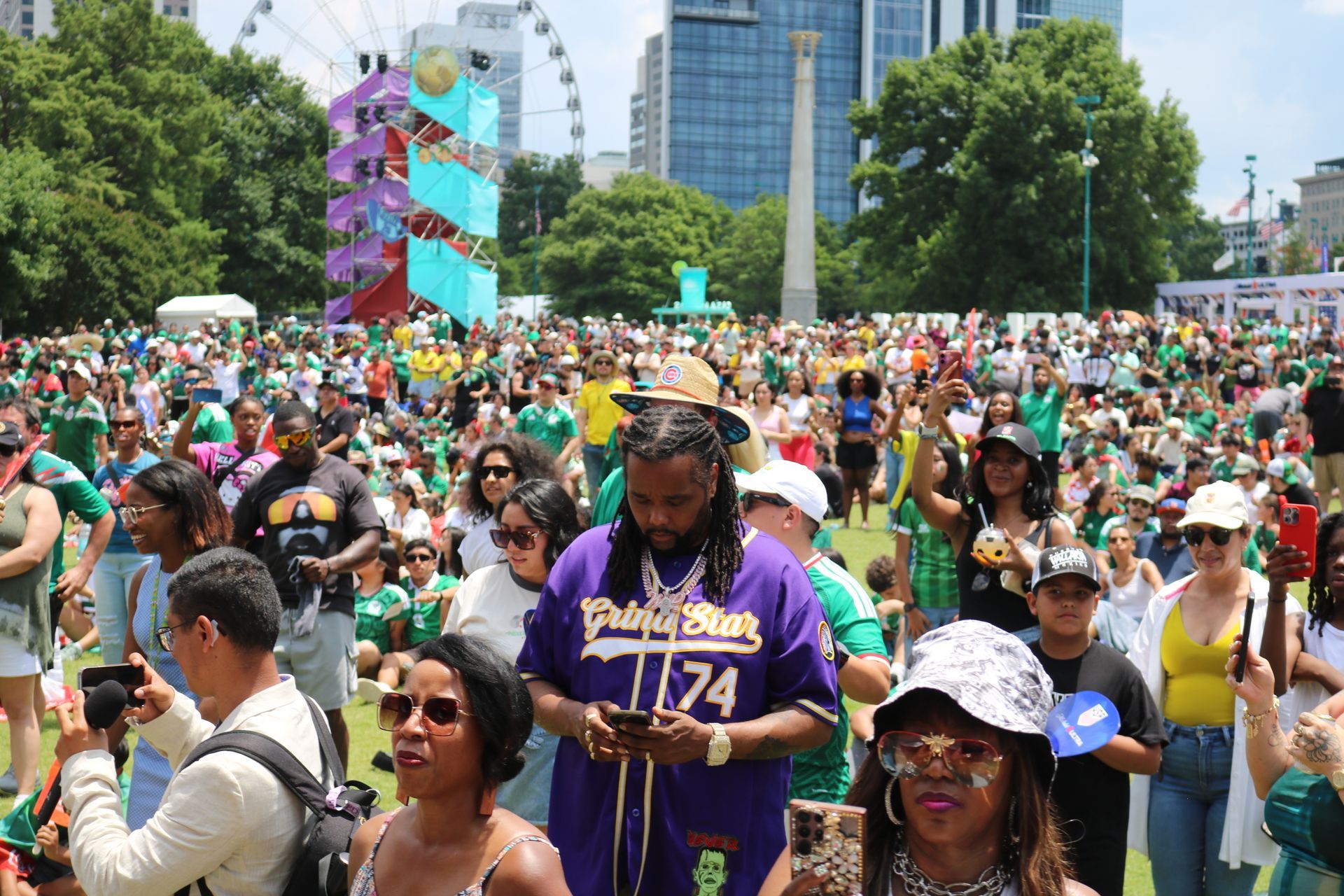 Crowd gathered at a colorful outdoor festival in a city park, with buildings and a Ferris wheel in the background