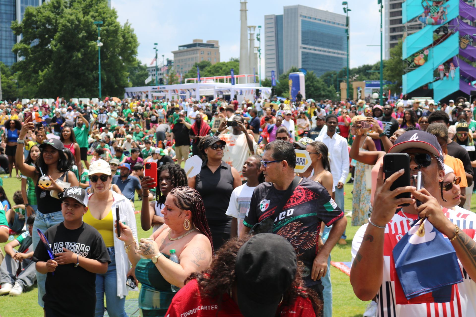 Crowd gathered at an outdoor city festival in bright sunlight, with people taking photos and buildings in the background