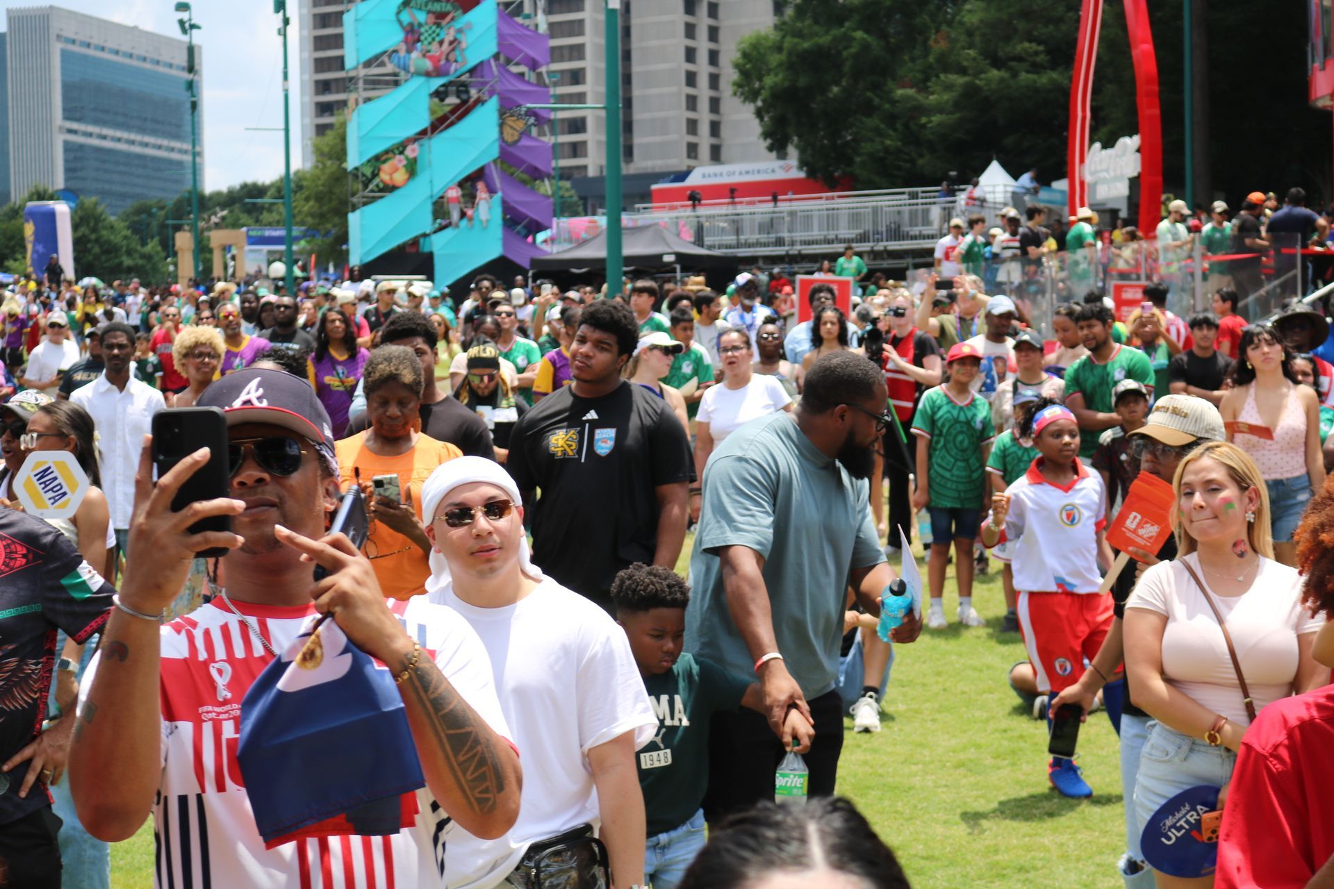 Crowd gathered on a grassy plaza at an outdoor festival, with people taking photos and colorful flags in the background.