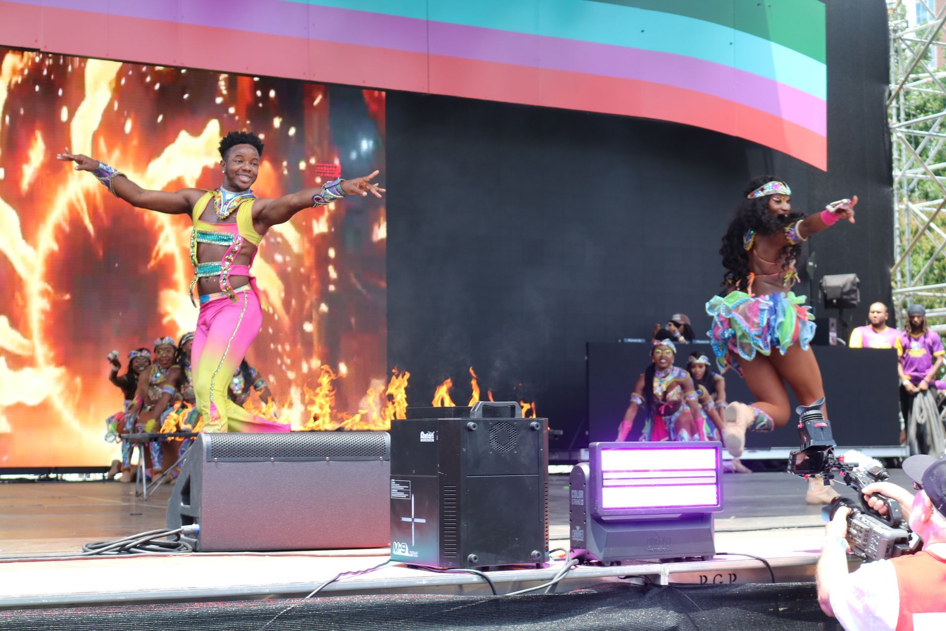 Two dancers in colorful costumes perform on an outdoor stage with bright LED flames and lights.