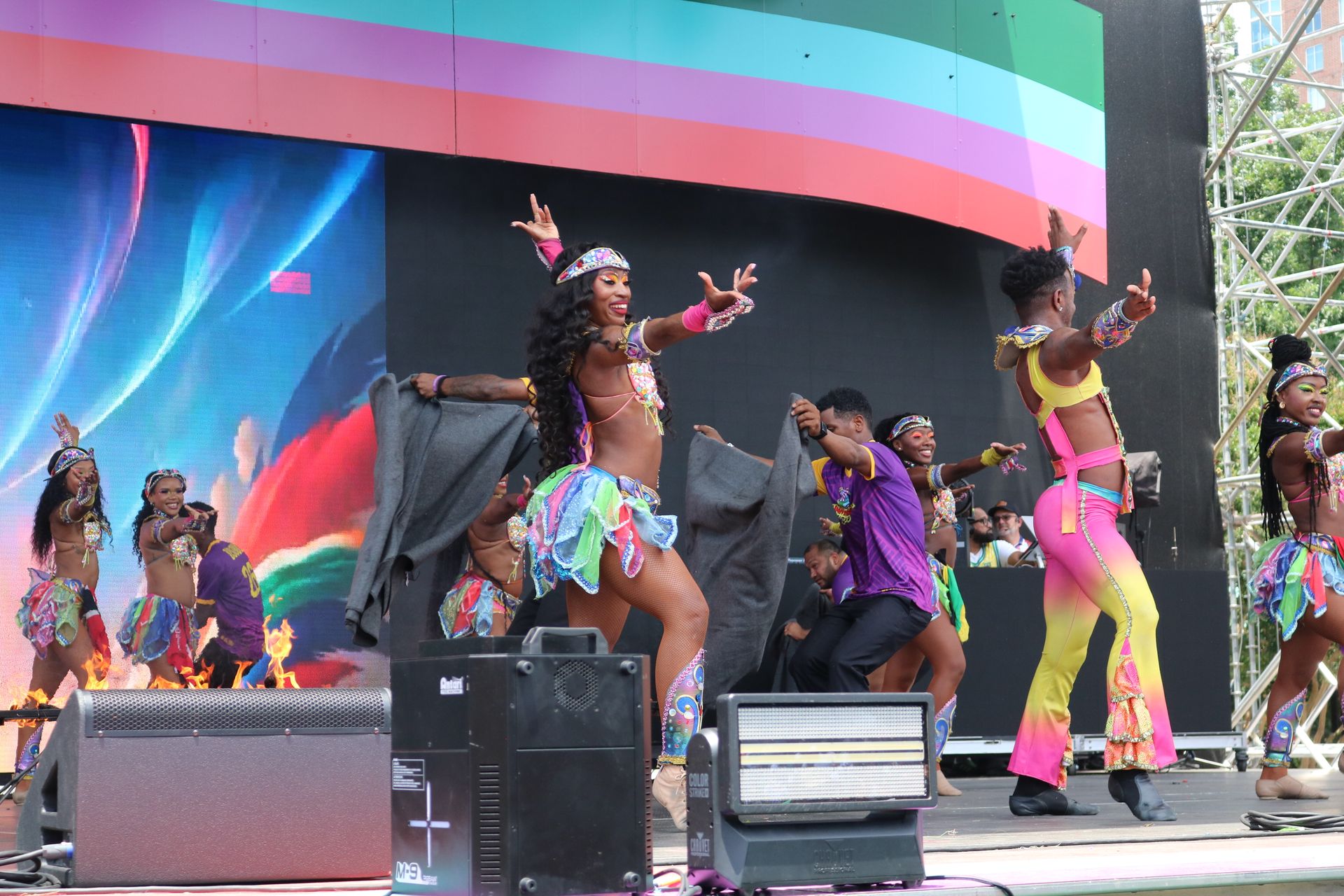 Performers dancing on an outdoor stage with colorful costumes and a rainbow backdrop