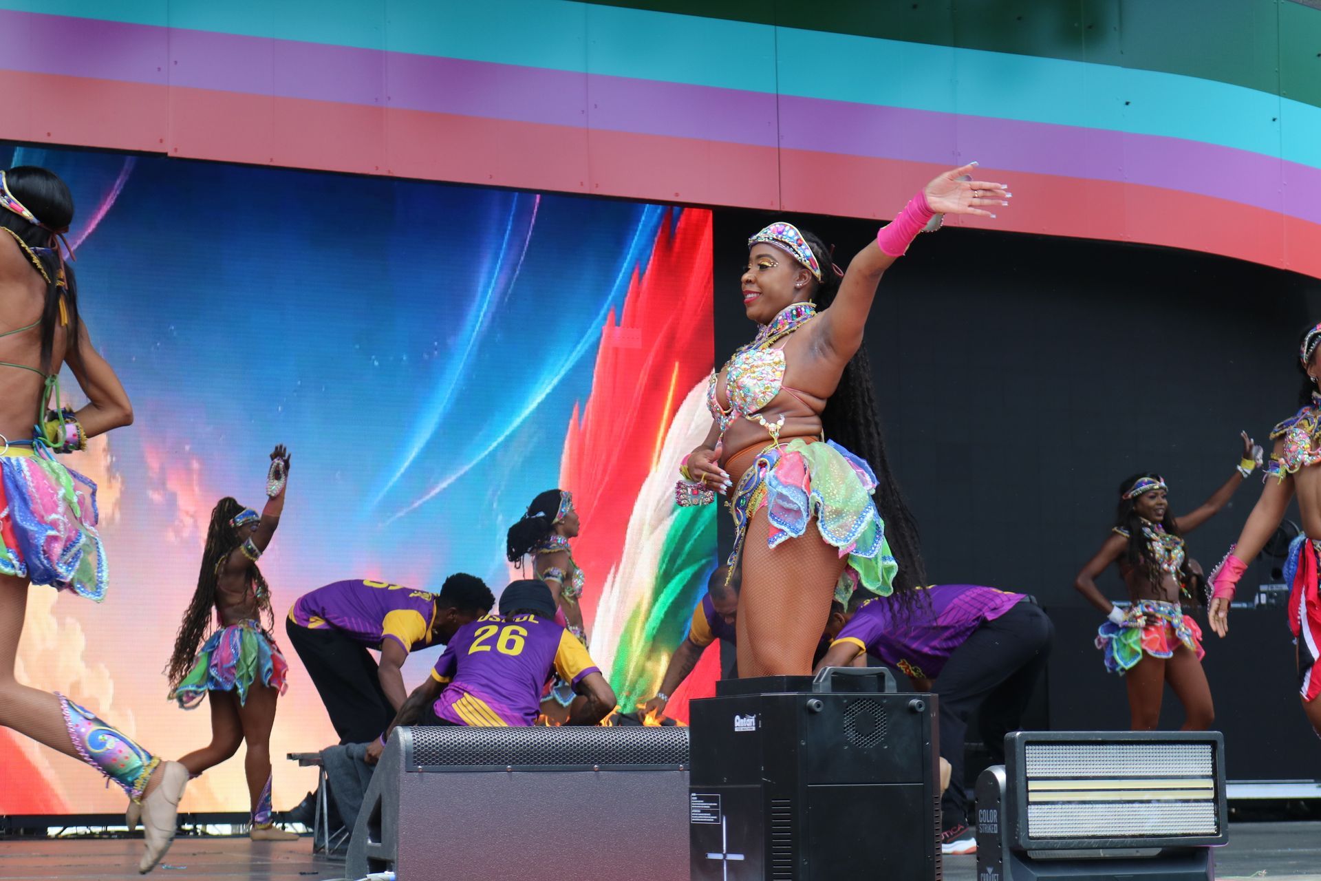 Dancers performing on a colorful outdoor stage with rainbow backdrops and black speakers in front