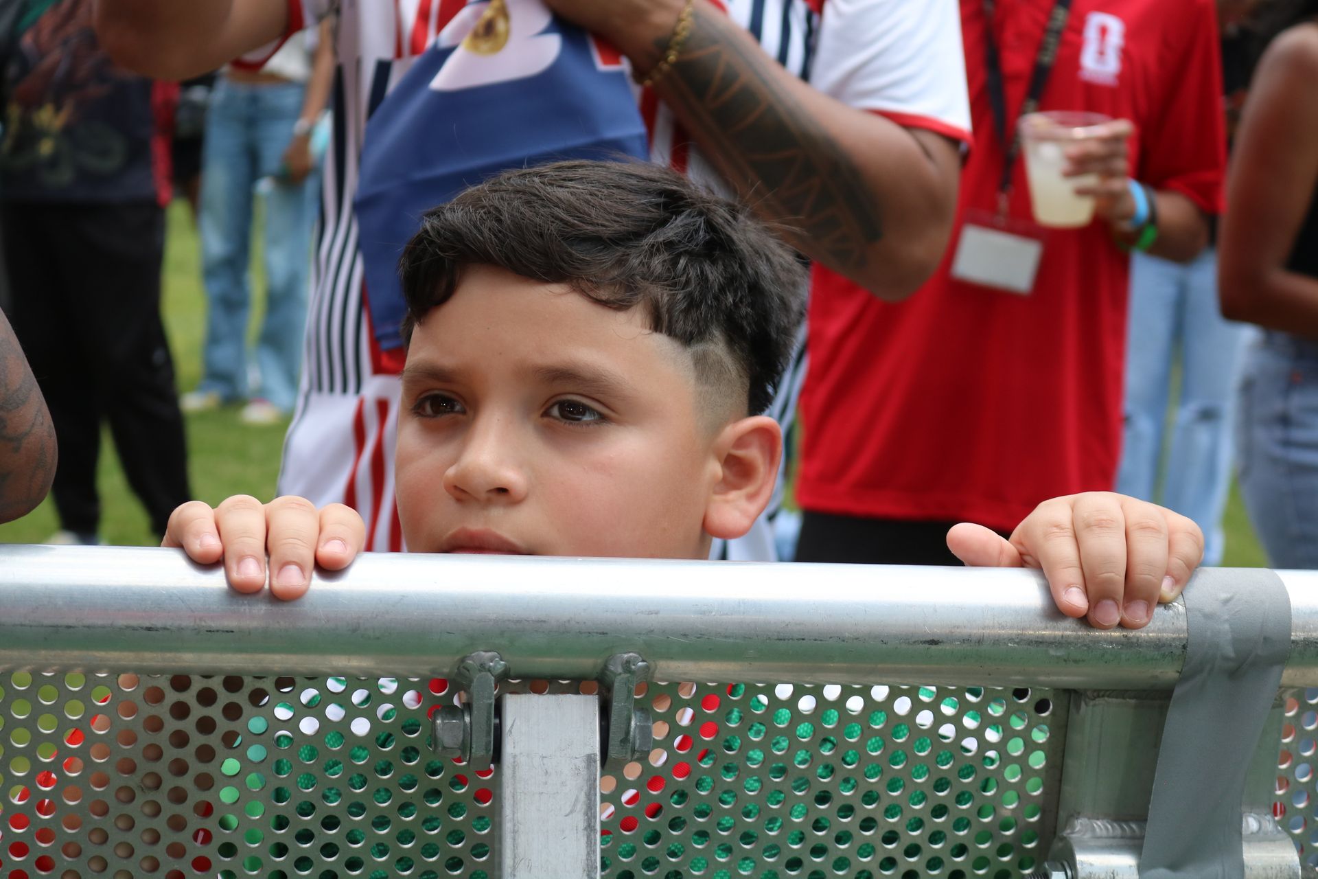 Boy peeking over a metal barricade at an outdoor event, with people standing behind him