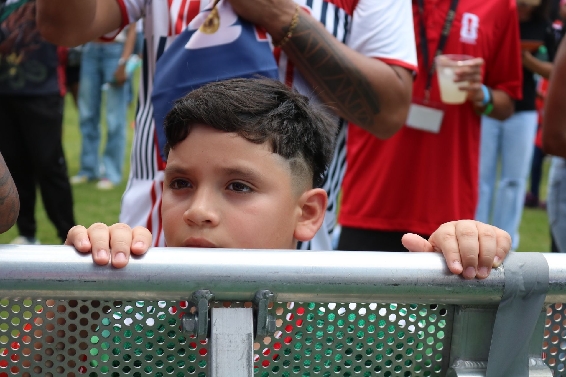 Child holding a metal barrier at an outdoor event, looking ahead with a serious expression