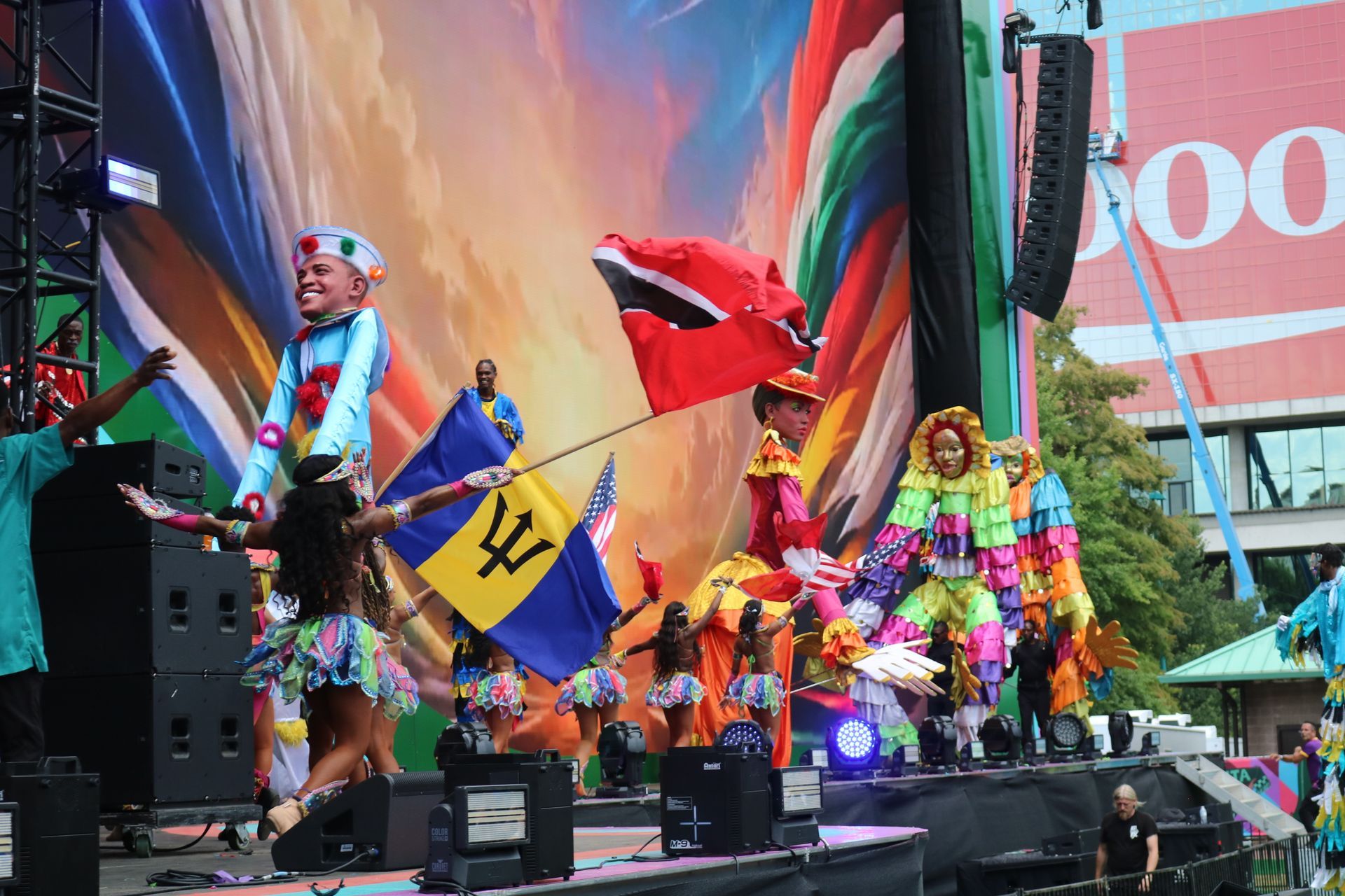 Performers in bright costumes dancing on a colorful outdoor stage with flags and speakers