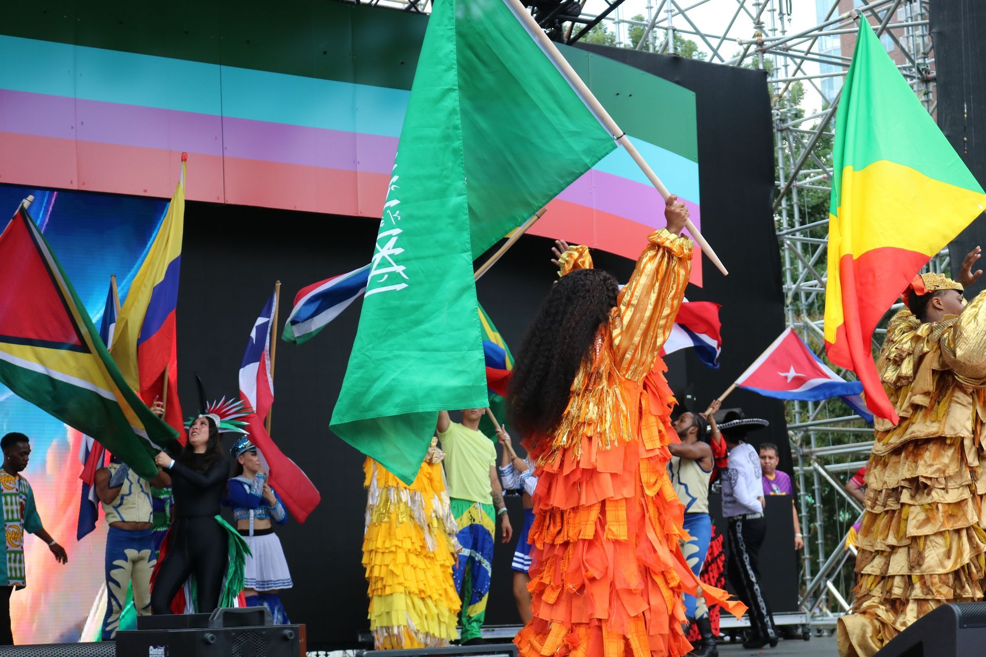 Dancers in colorful costumes wave large flags on an outdoor stage with a multicolored backdrop