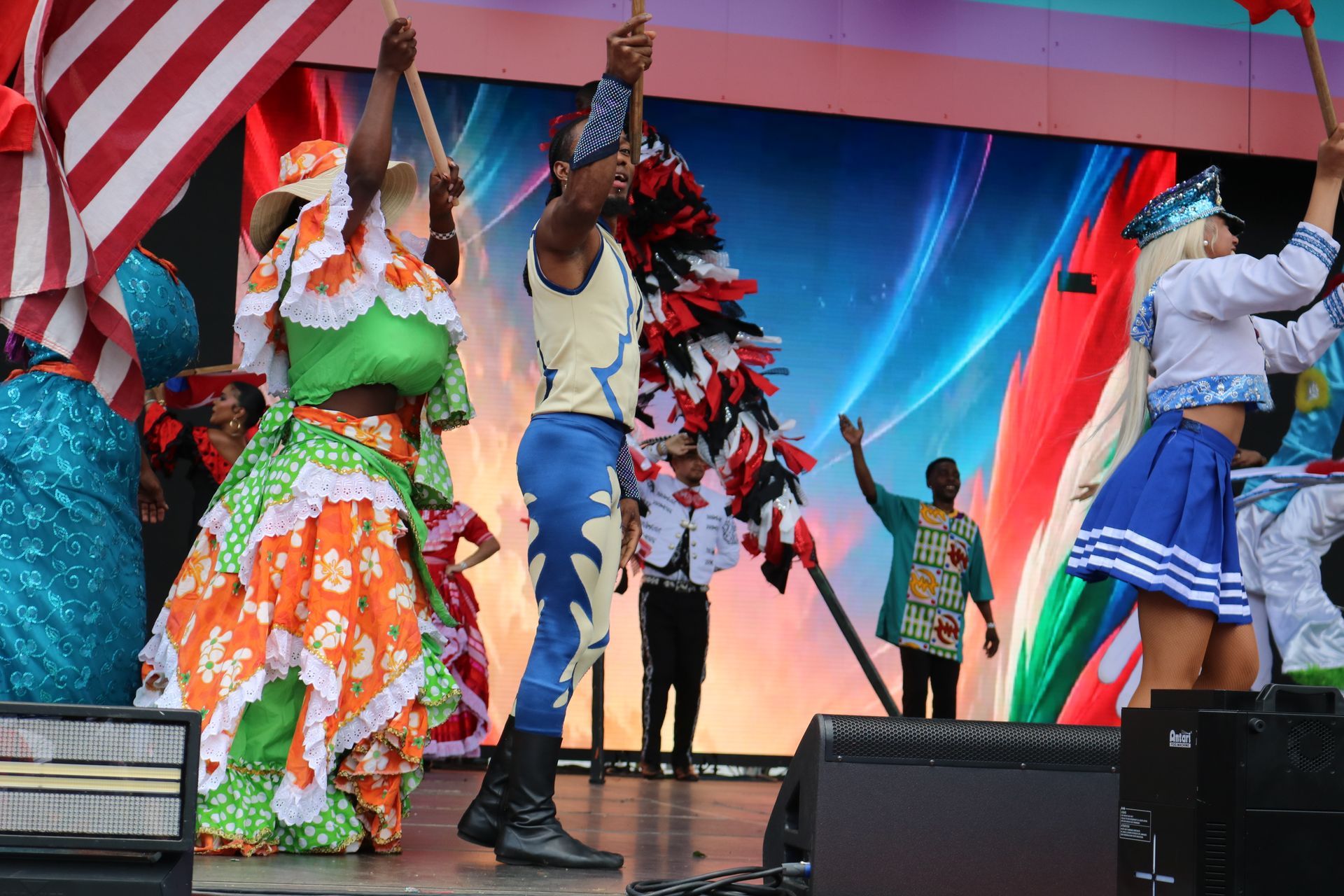 Performers in colorful costumes dancing onstage with red-and-blue drapes and a sunset backdrop