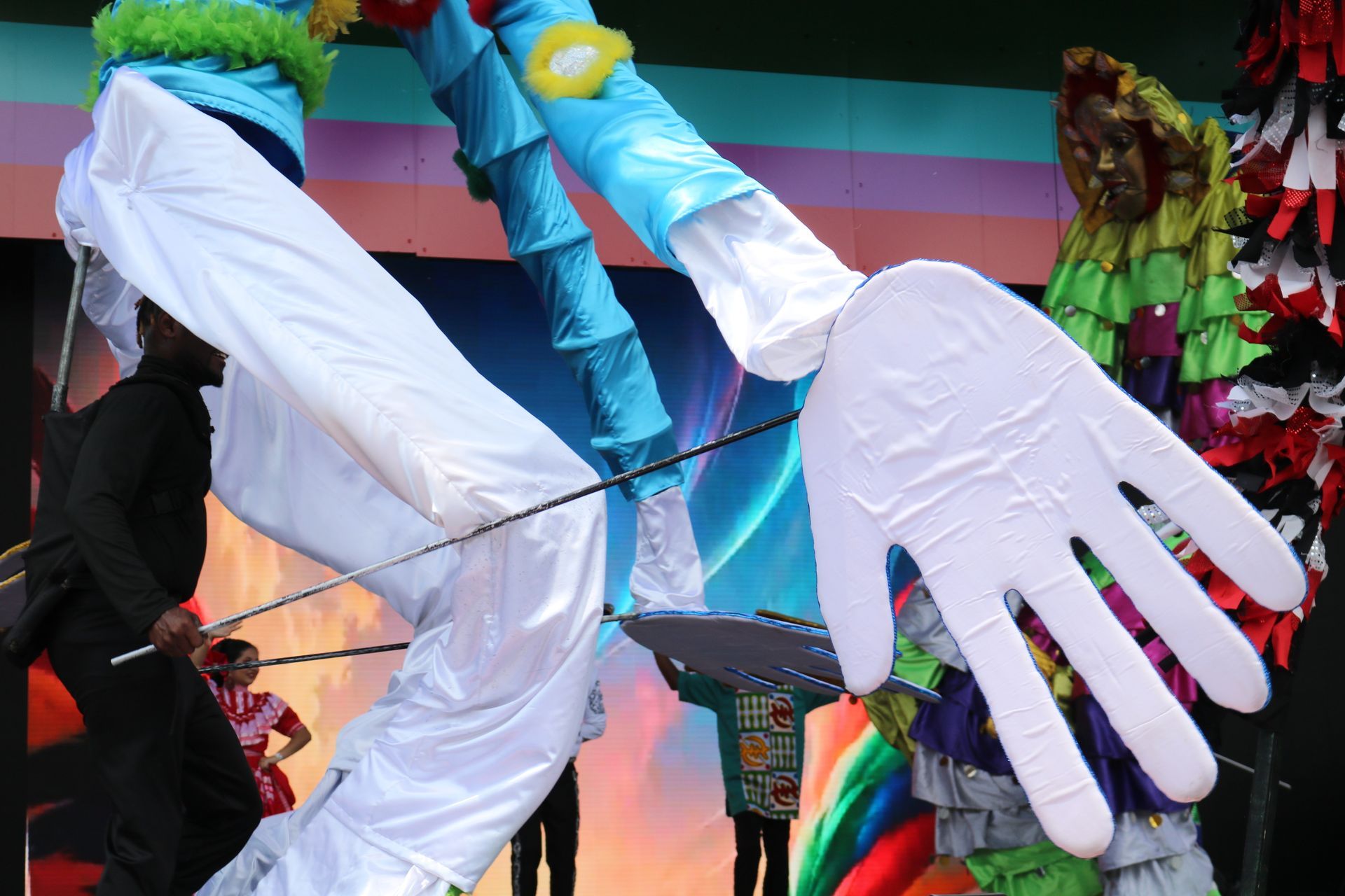 Person in white outfit with oversized white gloves and colorful feathers at a street parade