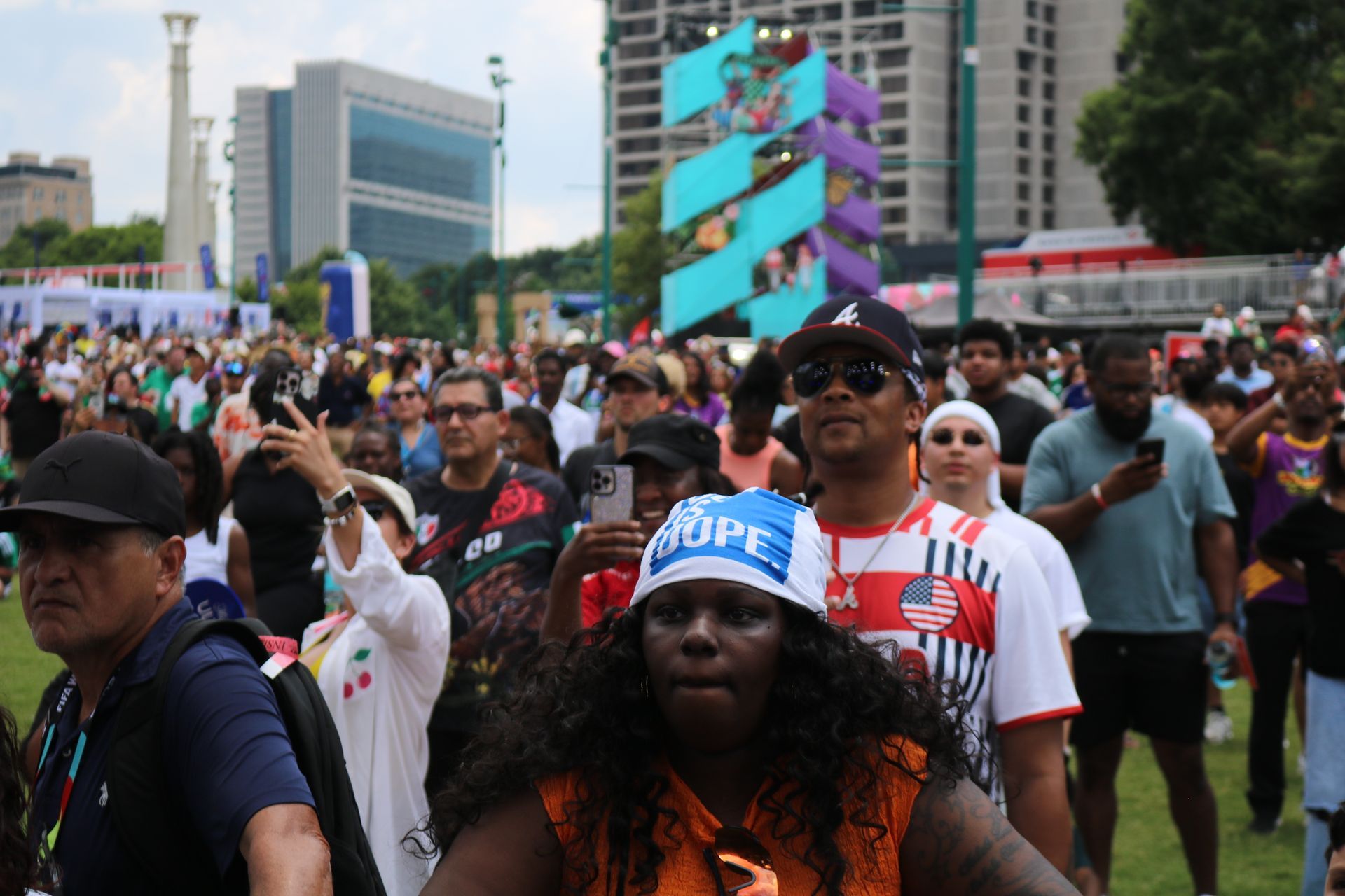 Crowd marching at an outdoor city rally, with people holding signs and a colorful sculpture in the background
