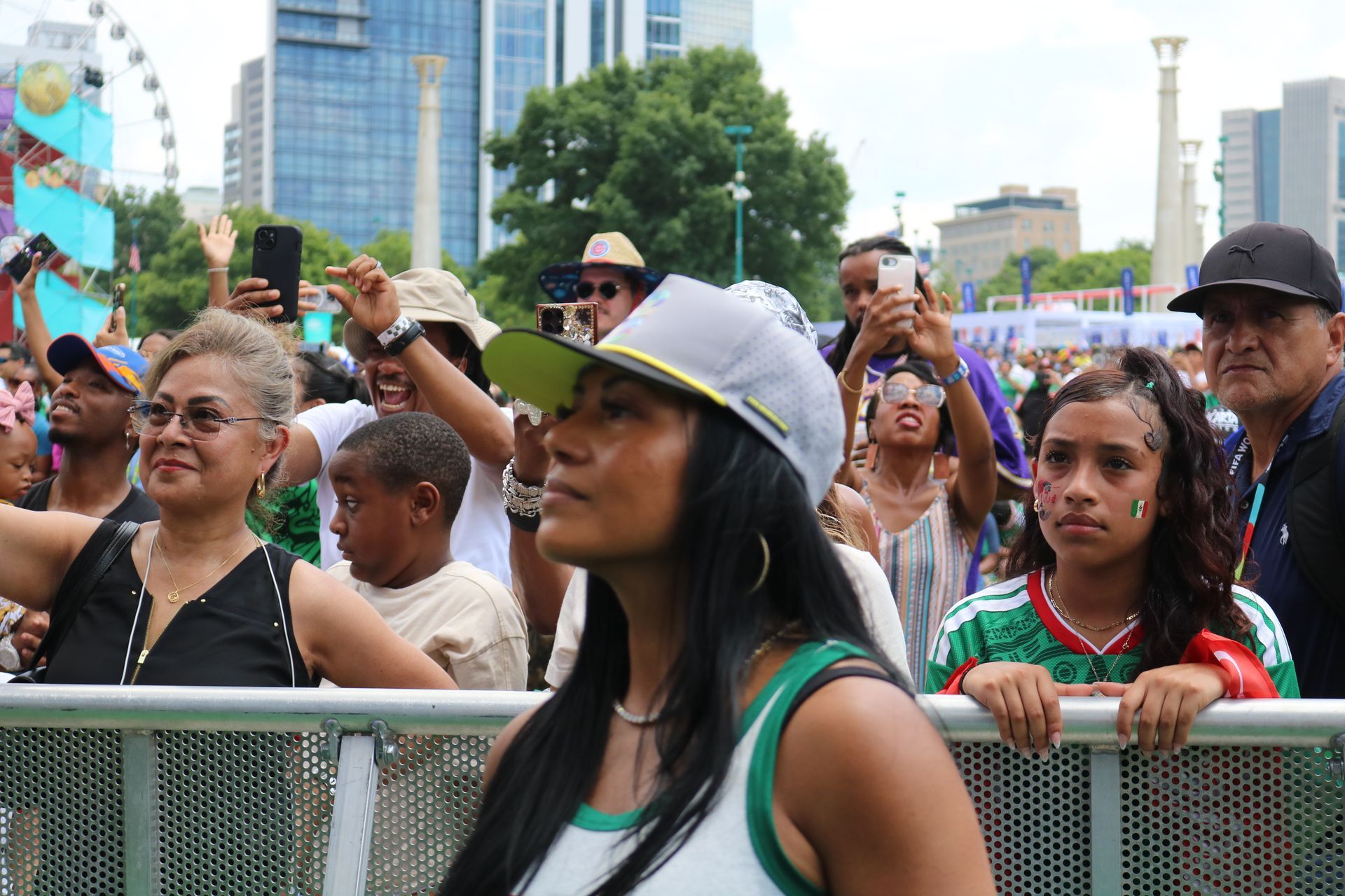 Crowd behind a metal barrier at an outdoor festival, watching and raising phones in the air