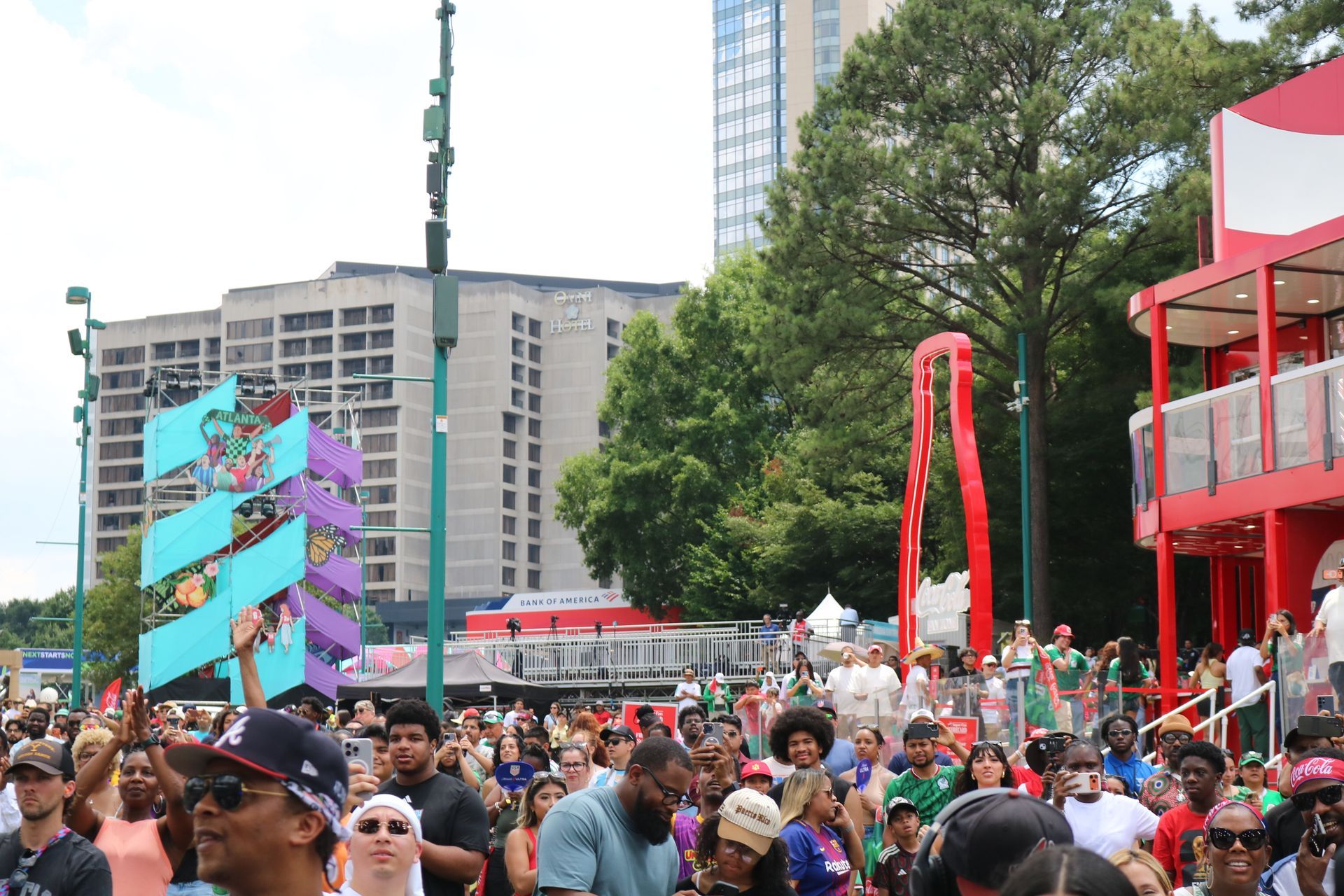 Crowded outdoor festival with colorful art structures, red stage, and tall buildings in the background