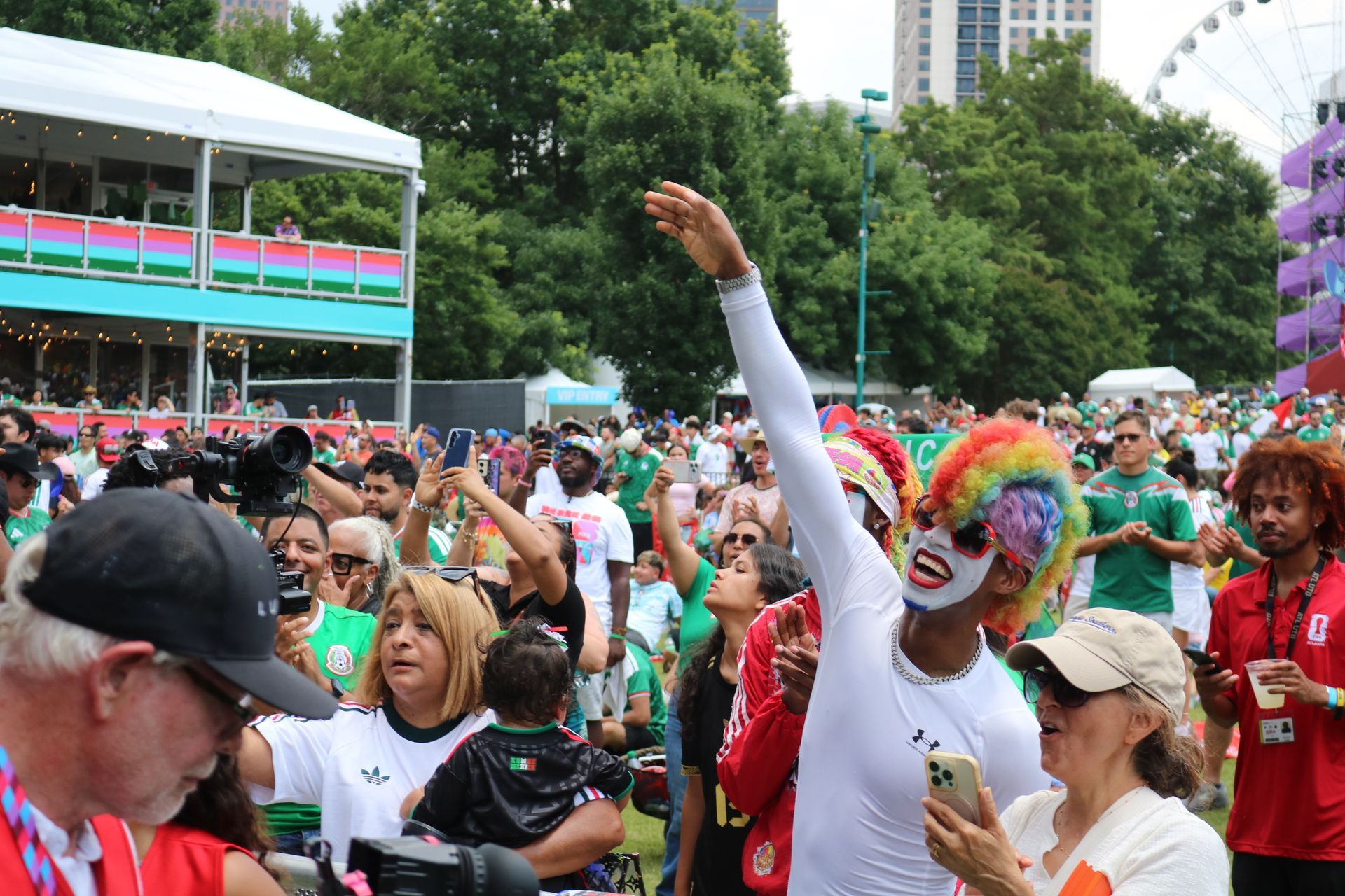 Crowd at a colorful outdoor parade, with a clown waving amid cheering spectators and a Ferris wheel behind.