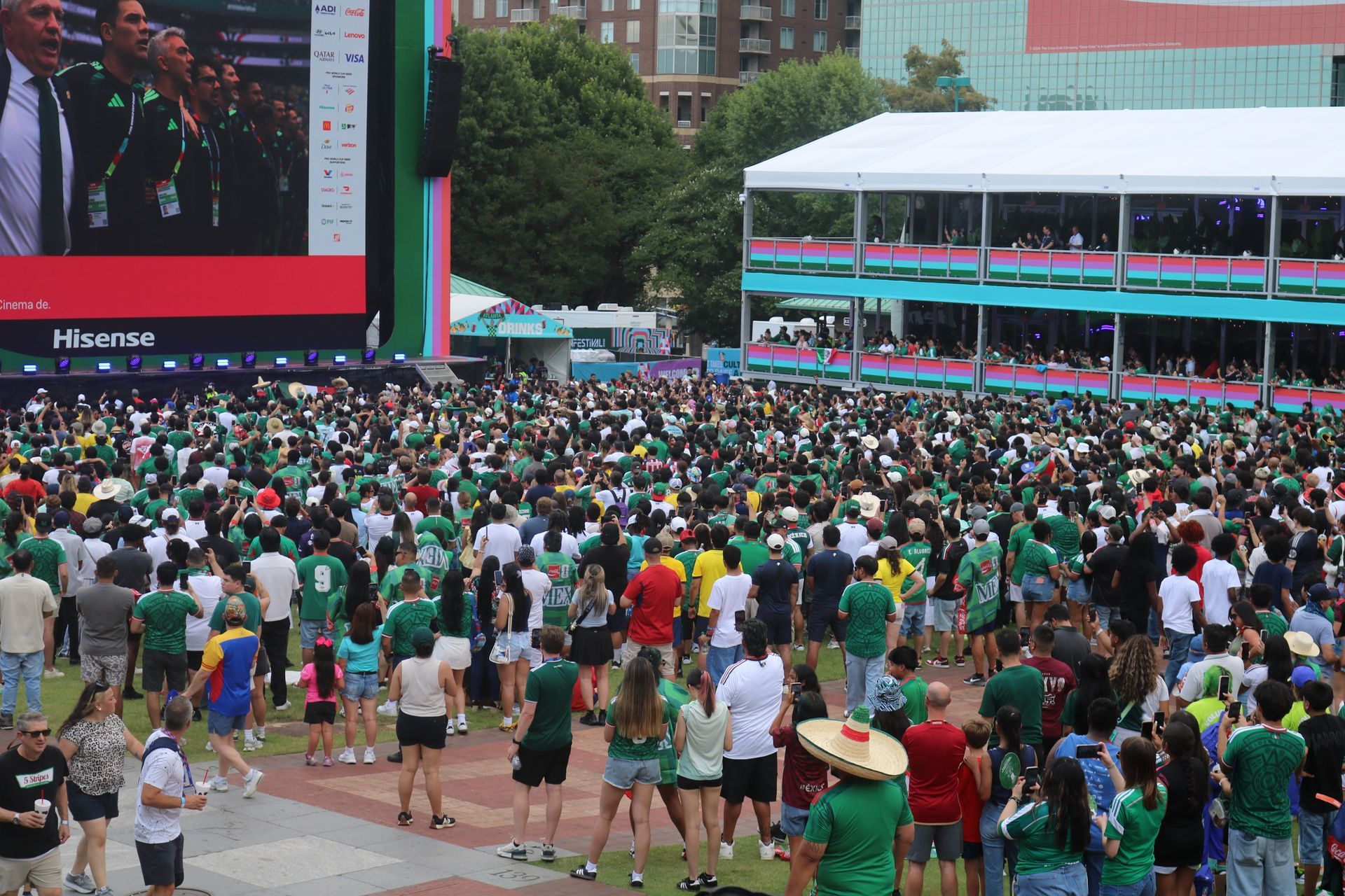 Crowd gathered in a stadium plaza watching a large screen during an outdoor event