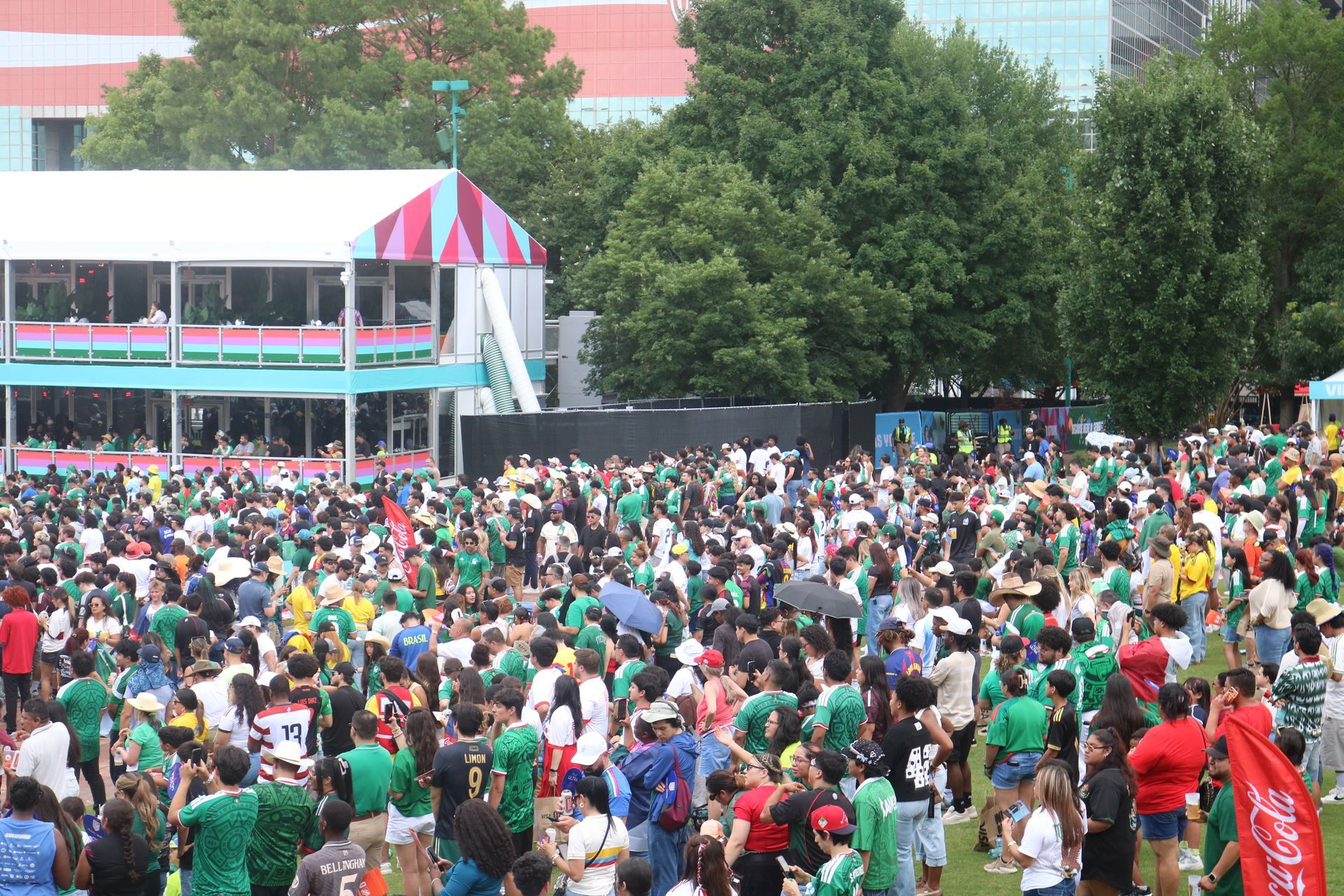 Large crowd gathered outdoors near a building with trees and red-and-white tents.