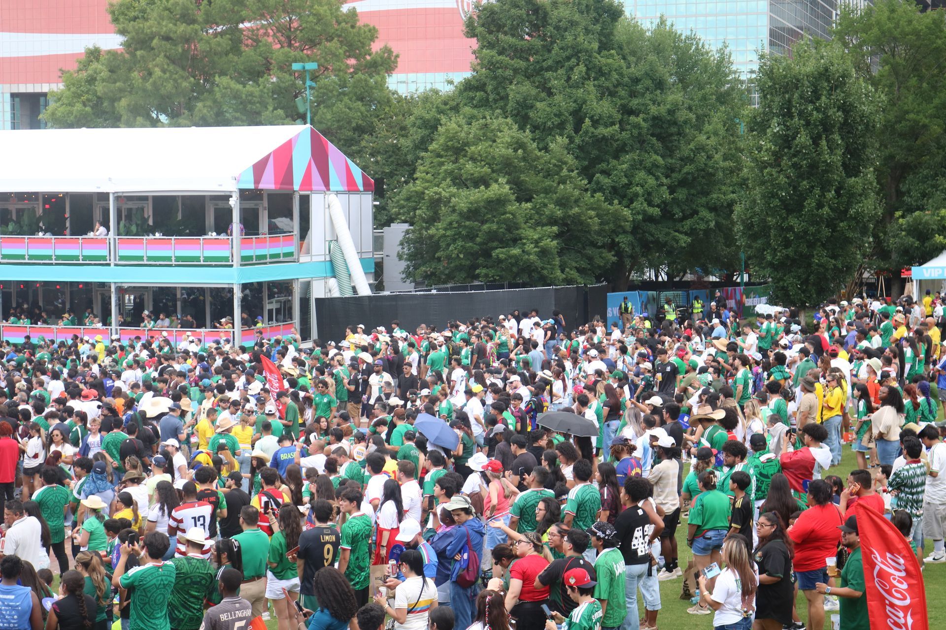 Crowd gathered outdoors near a colorful pavilion and trees, with people in green and white shirts.