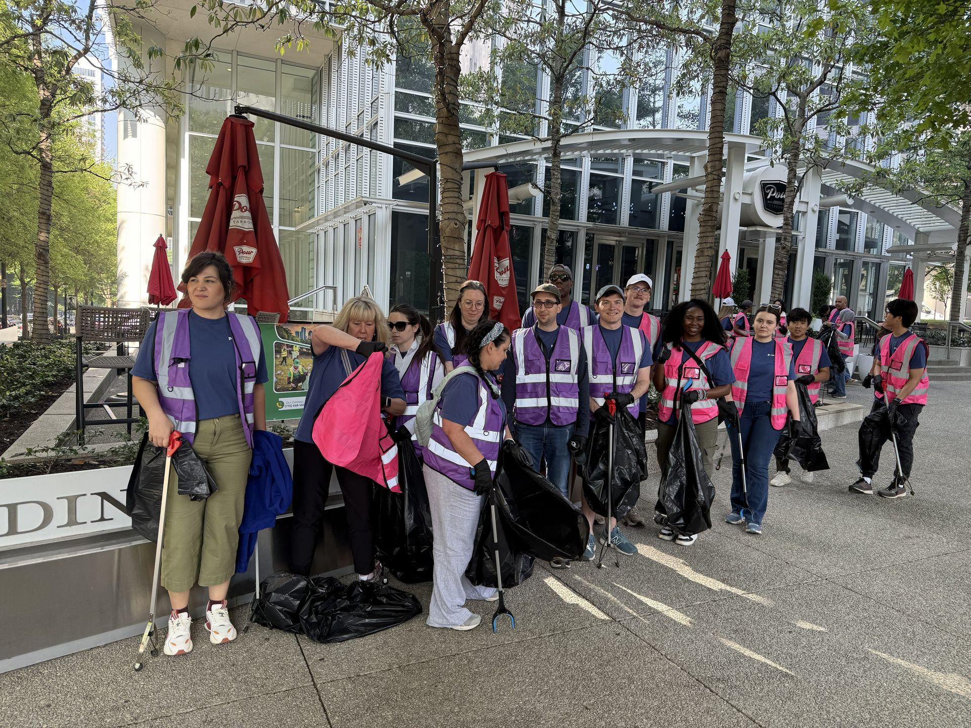 Group of people in purple vests and pink scarves standing outdoors near a building, holding red banners.