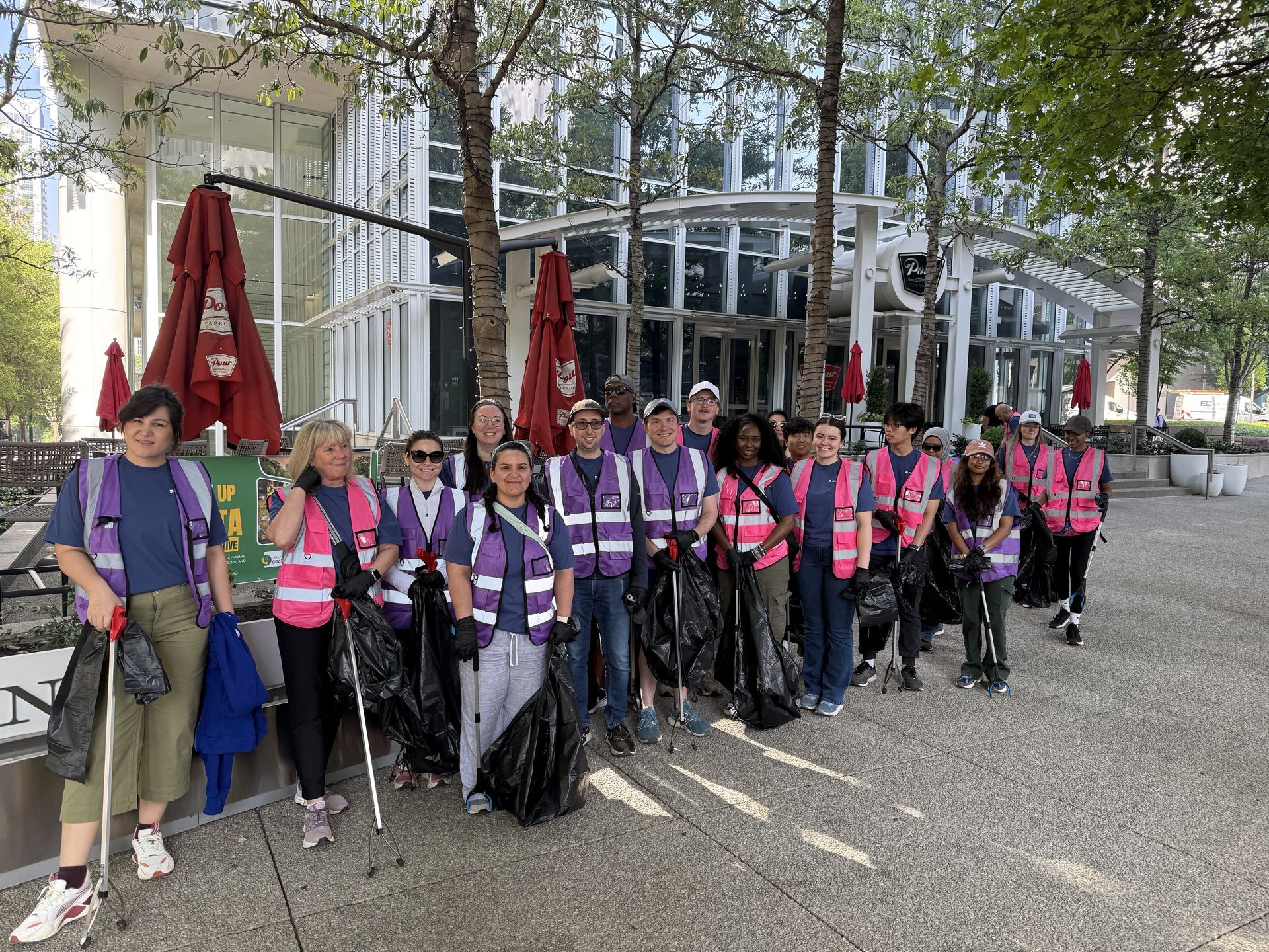 Group of volunteers in pink vests standing outdoors with bags and trekking poles near a building